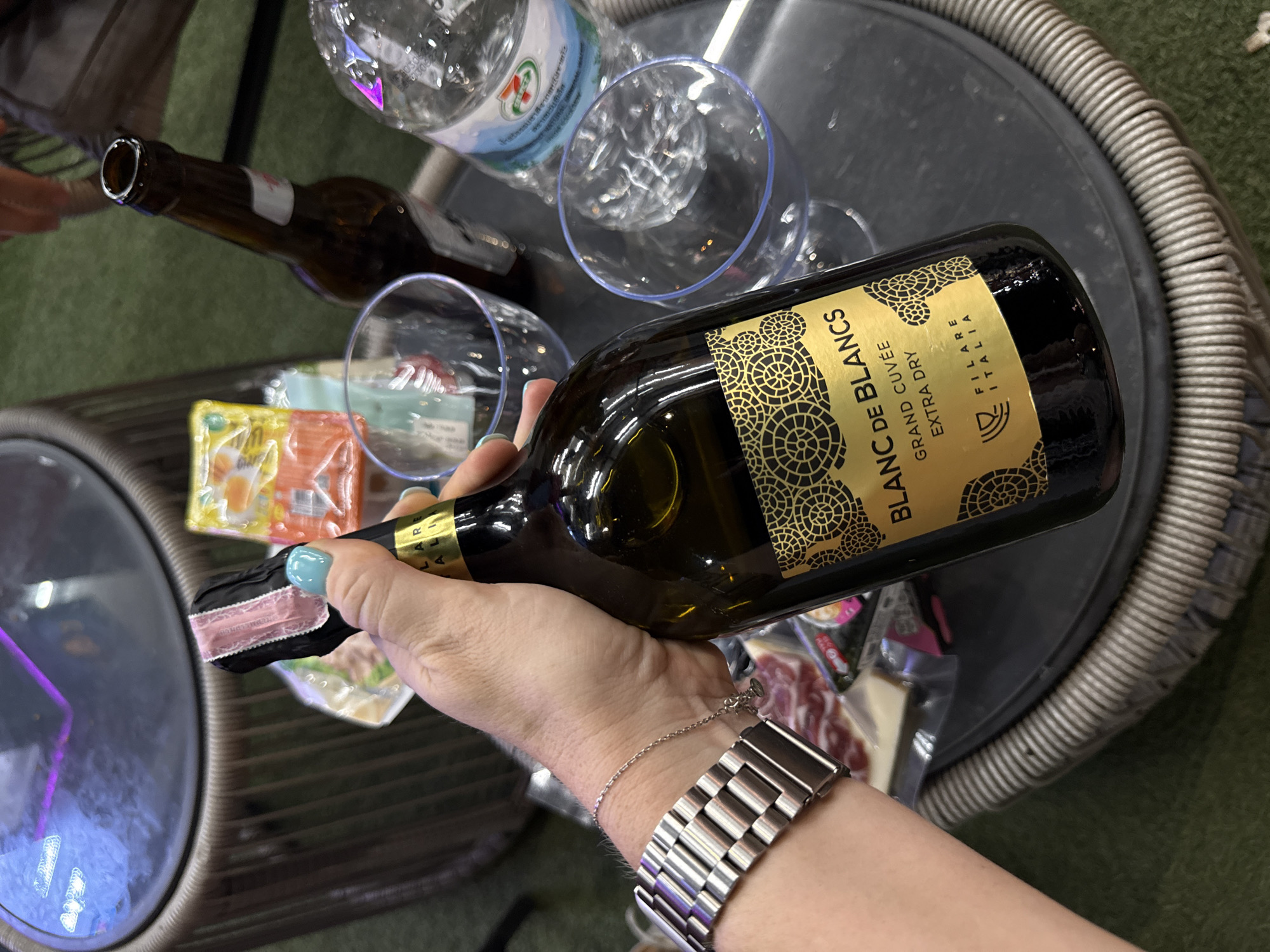 A person's hand holds a bottle of "BLANC DE BLANCS GRAND CUVÉE EXTRA DRY FILARE ITALIA" wine. The bottle has a gold label with a decorative pattern. The hand is wearing a silver watch and has light blue nail polish. In the background, there are two empty clear glasses, a bottle of water with a "7-ELEVEN" label, and what appears to be some food items in plastic packaging. The scene is set on a woven table, with a green surface that could be artificial turf or grass. The lighting suggests it could be daytime. The overall impression is a casual gathering or outdoor setting.
