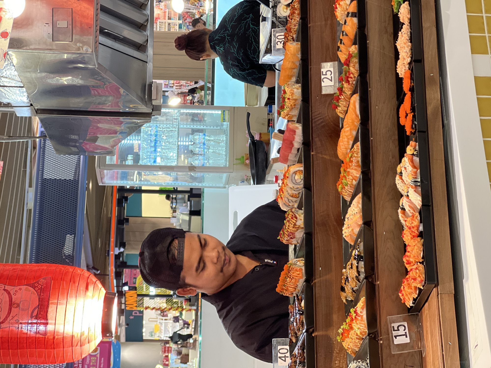 A medium shot captures a man and a woman behind a sushi bar in what appears to be an Unknown, Unknown, food market. The man, wearing a black cap and a black shirt with "PSUA" on it, is looking towards the sushi display. The woman, positioned further back, has her hair in a bun and is wearing a dark green shirt with a leaf pattern.
A variety of sushi is displayed on several tiers, each with a small price tag indicating the cost. The prices are "40", "15", "25", and "30".
The background reveals a busy food market. To the left, a red lantern illuminates the scene. Behind the sushi bar, refrigerated display cases can be seen, presumably holding drinks and other food items. Other stores or stalls can be seen in the distance.