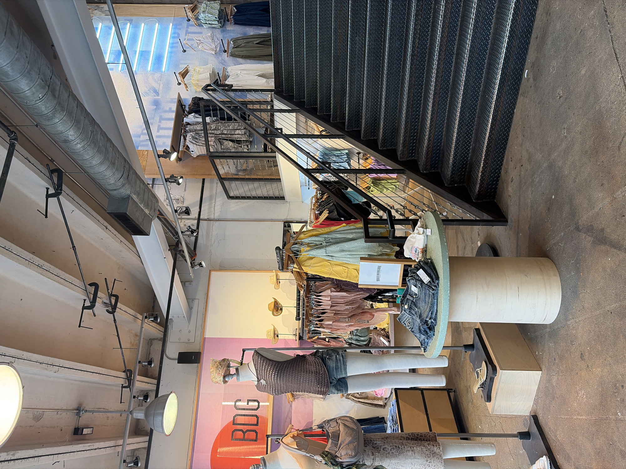 The image captures the interior of an Urban Outfitters store in Austin, Texas, USA. The scene showcases a clothing retail environment with garments displayed on mannequins and clothing racks. A prominent element is the staircase constructed of dark, industrial-style metal, leading to an upper level. The ceilings feature exposed ductwork and industrial lighting fixtures. The brand "BDG" is visible in large lettering, indicative of one of the clothing lines sold in the store. The interior design conveys an urban, contemporary aesthetic.