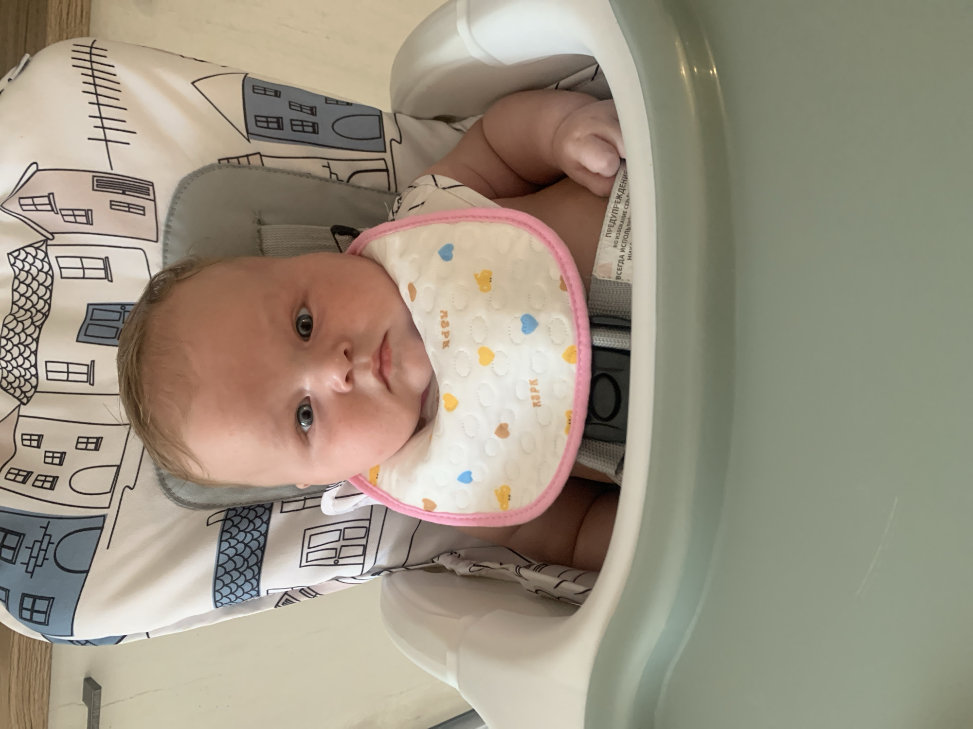A baby is lying in a high chair, wearing a bib with a pattern of small hearts and chicks. The bib has a pink trim. The high chair has a light gray cushion with a pattern of houses in black and blue. The baby has blue eyes and light brown hair. The baby is looking at the camera with a neutral expression. There is a warning label visible on the side of the high chair, with text in Russian that appears to read "ПРЕДУПРЕЖДЕНИЕ" and "ВСЕГДА ИСПОЛЬЗУЙТЕ СИДЕНЬЯ". The scene appears to be indoors, possibly in a home setting in Stavropol, Russia, as there is a wooden surface visible in the background. The lighting suggests it is daytime.