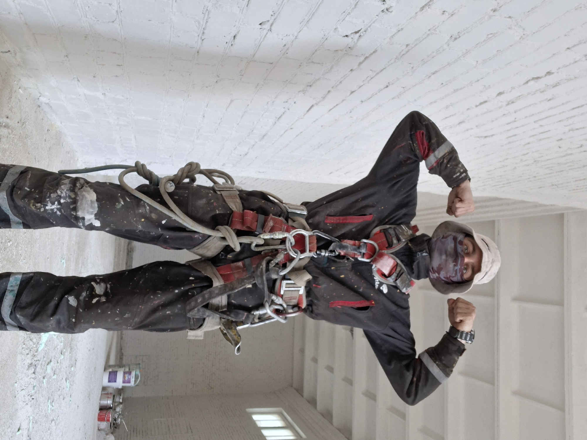 A man in a dark jumpsuit and safety harness stands indoors against a white-painted brick wall. He wears a light-colored hat and a mask obscuring the lower part of his face. Paint splatters are visible on his clothing. He is flexing his fists, showing a watch on his left wrist. The scene is lit with bright indoor lighting. Paint cans are visible on the floor. The city is Unknown, Unknown.