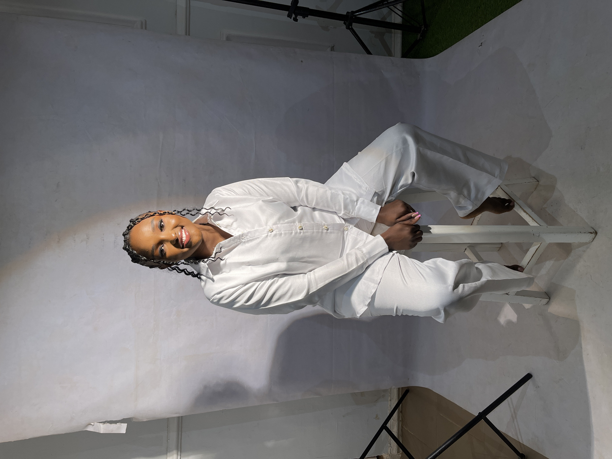 A young Black woman with braided hair and a bright smile is sitting on a white stool in a studio. She is wearing a white button-down shirt and matching white pants. The background is a textured white paper backdrop, with a spotlight creating a shadow on the left side of the image. To the left of the woman, a black tripod is visible, and to the right, another tripod with a light fixture is partially visible. The floor is a light gray color, with shadows cast from the stool and the woman. The overall impression is a professional studio portrait. The mention of Barakin Akawo, Nigeria, does not correspond to any visual information present in the image.