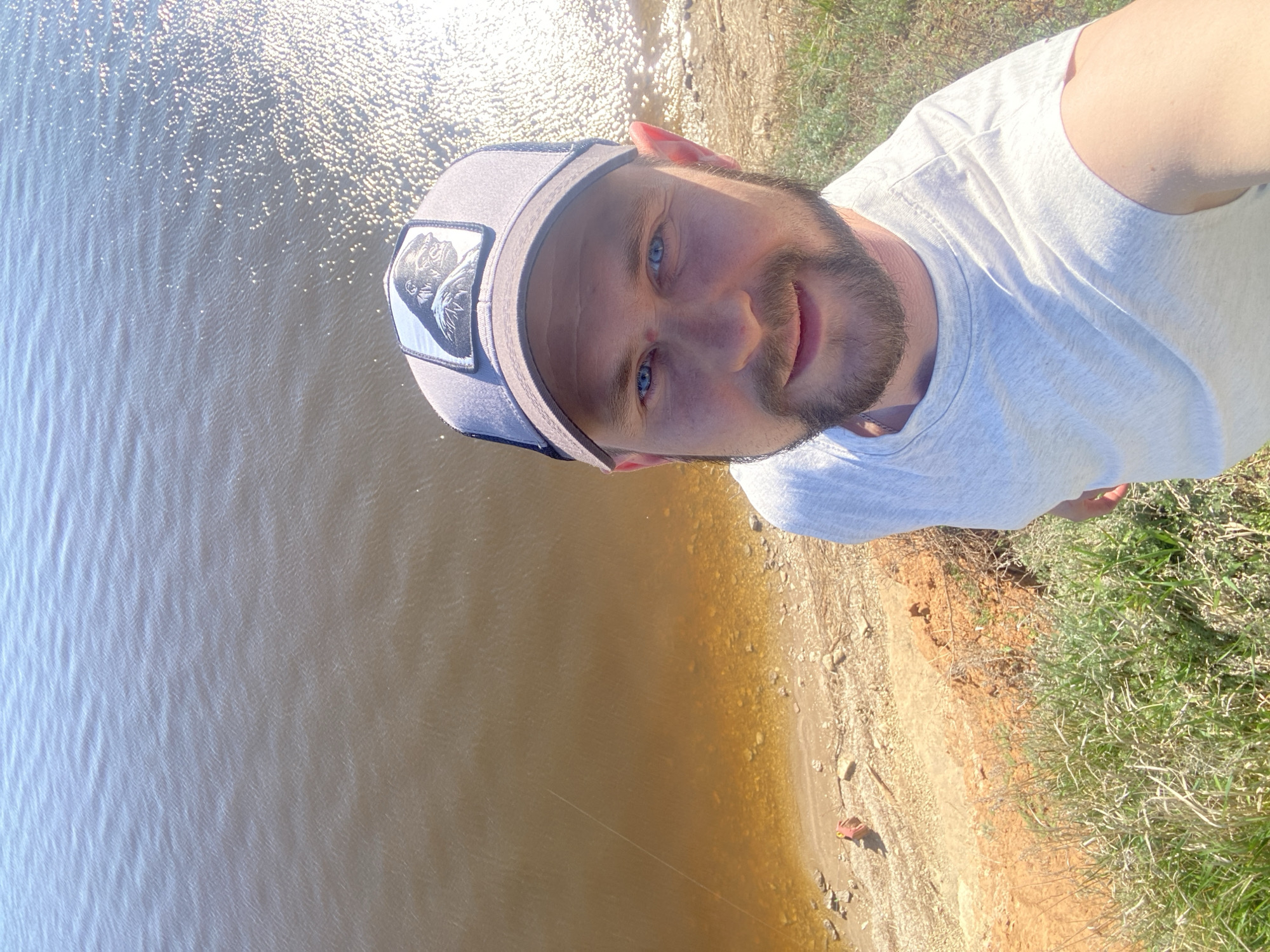 A selfie depicts a man in a gray cap with a gorilla patch and a white t-shirt, positioned near a body of water with ripples, and a shoreline transitioning from sand to grass. The location appears to be Шуран, Russia.