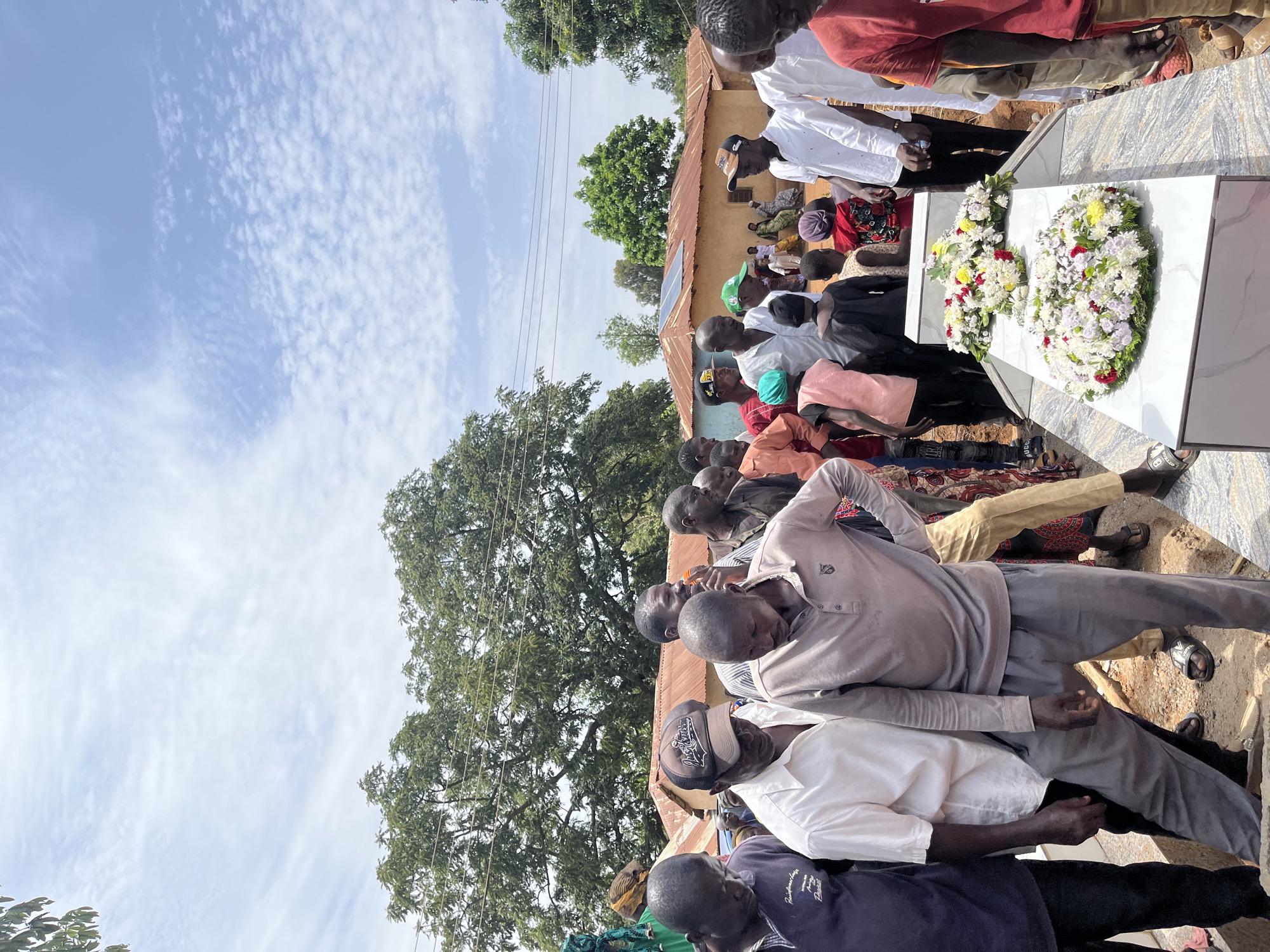The image shows a group of people gathered around a white grave covered in floral wreaths, likely attending a funeral in Dutsen Kato, Nigeria. Most of the people in attendance are men of African descent. The environment includes trees, a partly cloudy sky, and a nearby building with a corrugated metal roof and solar panels. The attendees are standing, some with their heads bowed, suggesting a moment of mourning or respect.