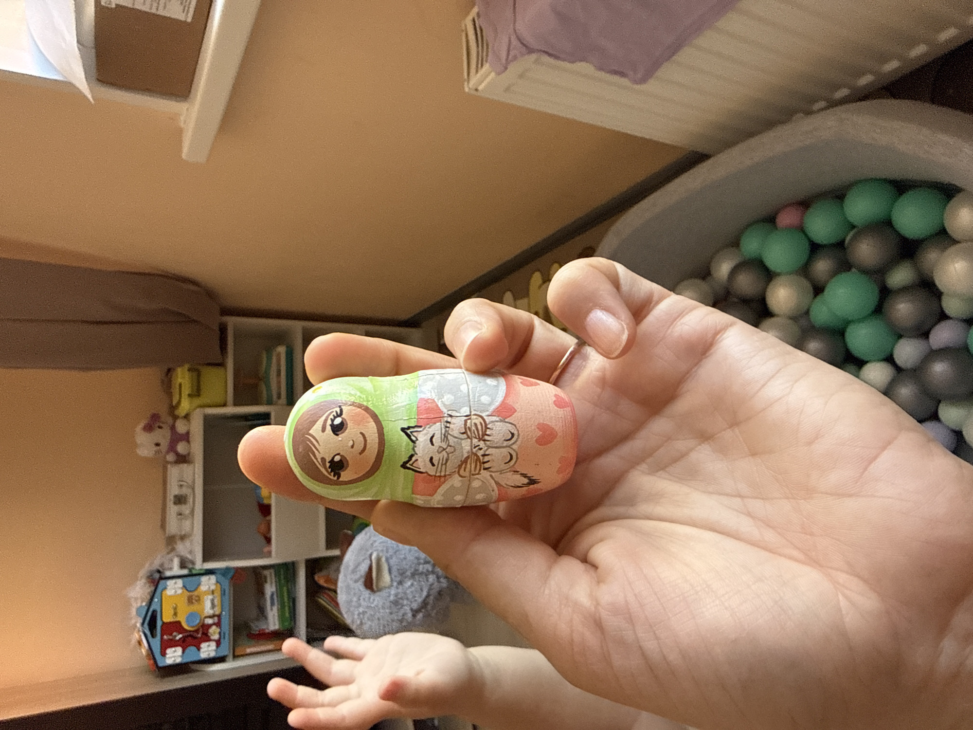 An indoor, slightly rotated image captures a domestic scene, likely a child's playroom or bedroom, in Tobolsk, Russia. The primary focus is a hand, presumably an adult's, holding a decorative toy resembling a Matryoshka doll. The toy, appearing to be made of wood, is split into two halves; one end is predominantly green with the painted face of a smiling girl with brown hair, while the other end is pink, depicting a light-colored cat surrounded by small hearts. A simple ring is visible on the adult's ring finger.
In the background to the right, a grey ball pit is partially visible, filled with numerous plastic balls in shades of green, grey, and possibly light pink or lavender. To the left, a child's hands and arms are discernible, reaching out, suggesting interaction or play. Behind the child, a grey plush toy is partly obscured.
Further in the background, a shelving unit is visible against a light brown wall, displaying various children's items. These include a yellow toy, a white Hello Kitty plush toy, a blue and red activity board or toy, and several books. The overall impression is a vibrant and active child-friendly environment, illuminated by ambient daylight. No discernible text is clearly visible on any objects.