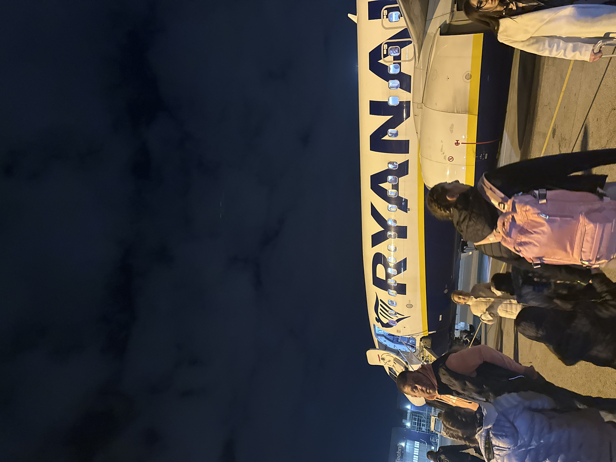 The image shows passengers disembarking from a Ryanair airplane at night. The side of the plane is visible, with the airline's name prominently displayed. Several people are walking on the tarmac, seemingly towards the terminal. The building in the bottom of the photo has the word "Technik" on the side, and the setting is Sofia, Bulgaria.