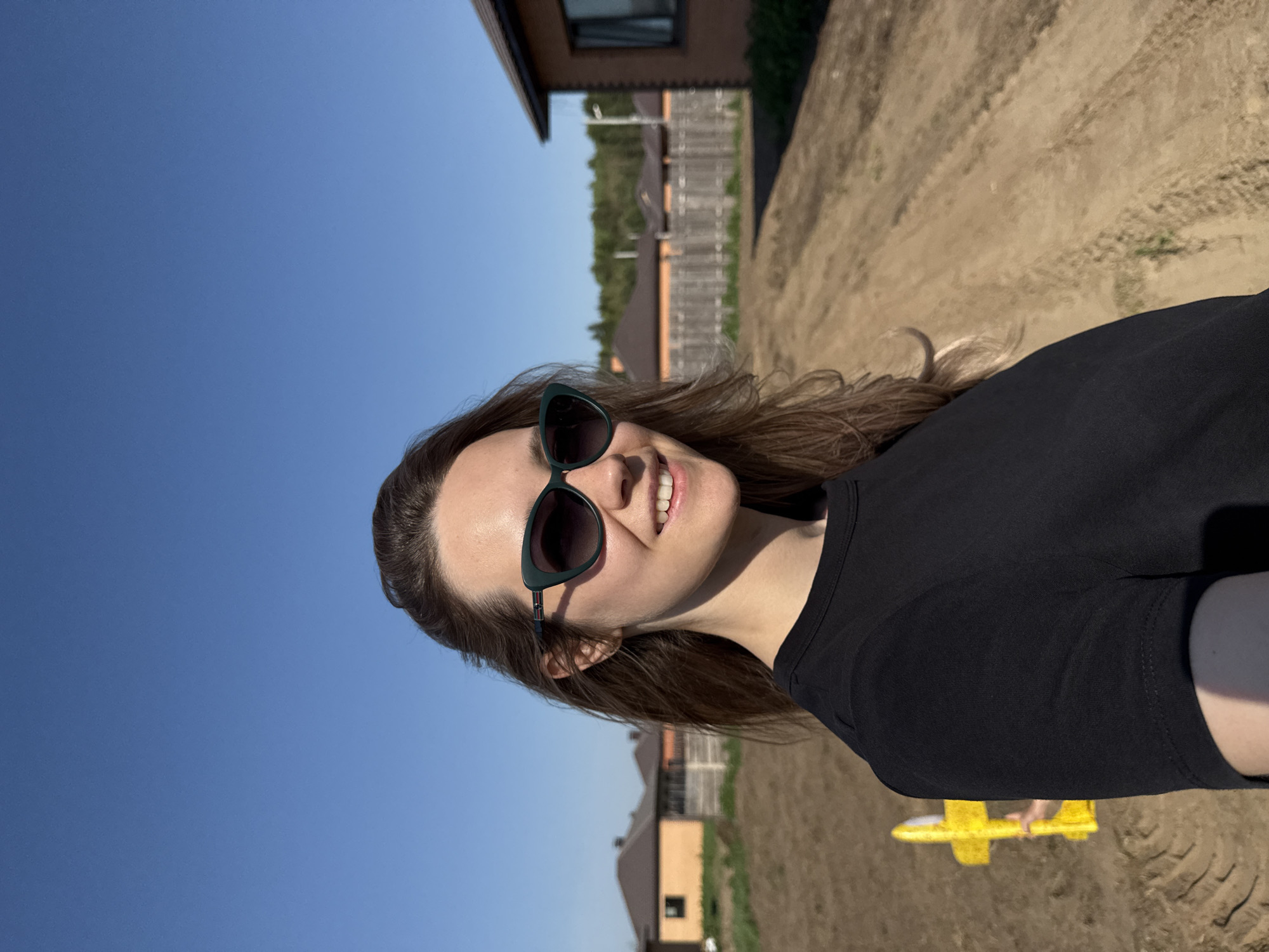 A woman with brown hair and sunglasses stands outdoors. She is wearing a black shirt. In the background, there are houses, fences, and a blue sky. A yellow object, possibly a toy, is at the bottom of the frame. The location is Chaikovsky, Russia.