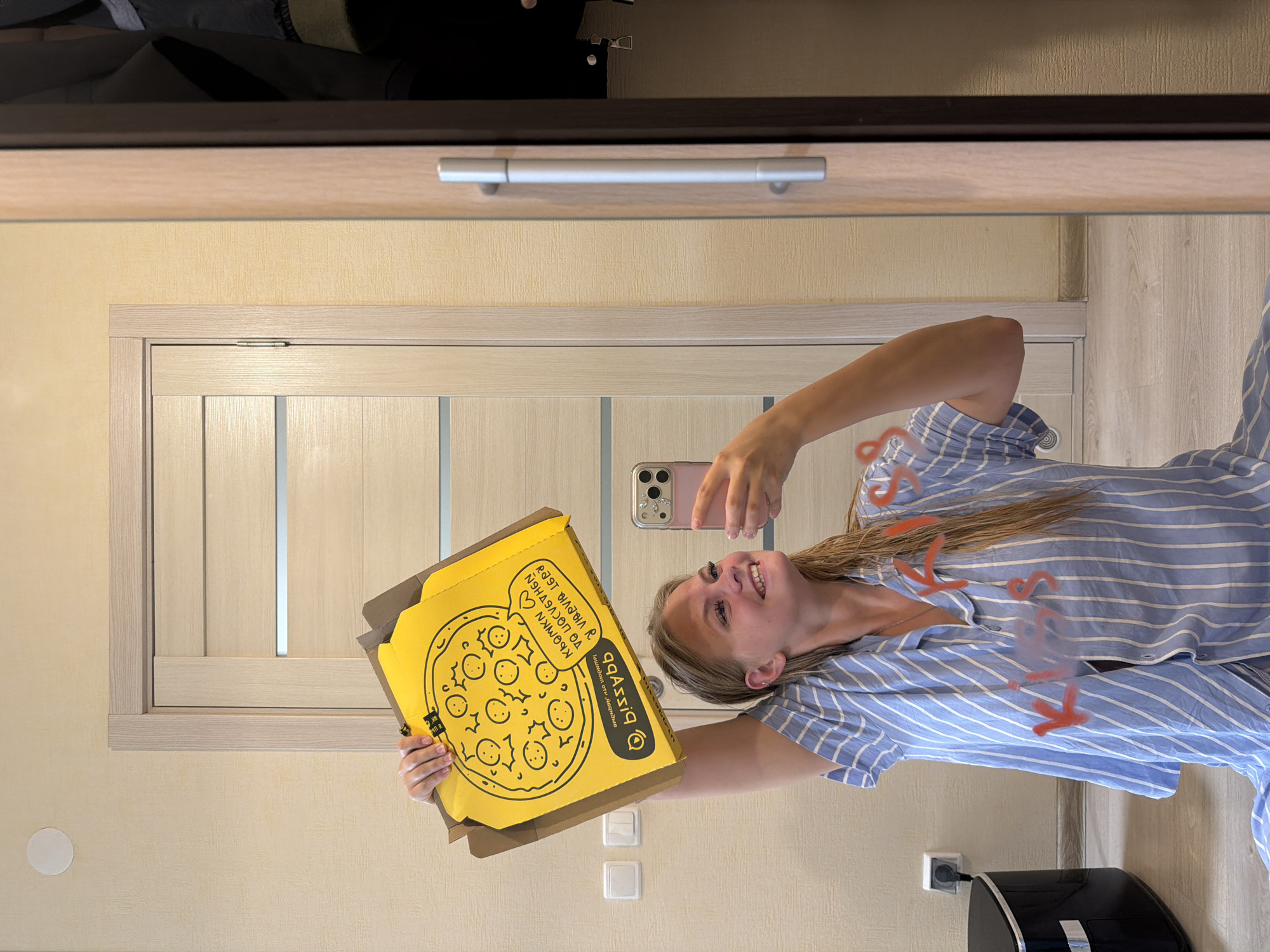 A young woman with blonde hair, wearing a blue and white striped pajama shirt, is taking a selfie in a mirror. She is holding a yellow pizza box with a cartoonish pizza design and text in what appears to be Russian. Her expression is joyful. In the background, a light-colored wooden door is visible, and the room has beige textured walls. The scene appears to be indoors, possibly a bedroom or hallway, and the lighting suggests it could be daytime. The woman is holding her phone up to take the photo, and the reflection shows her holding the pizza box. The image is oriented vertically.
