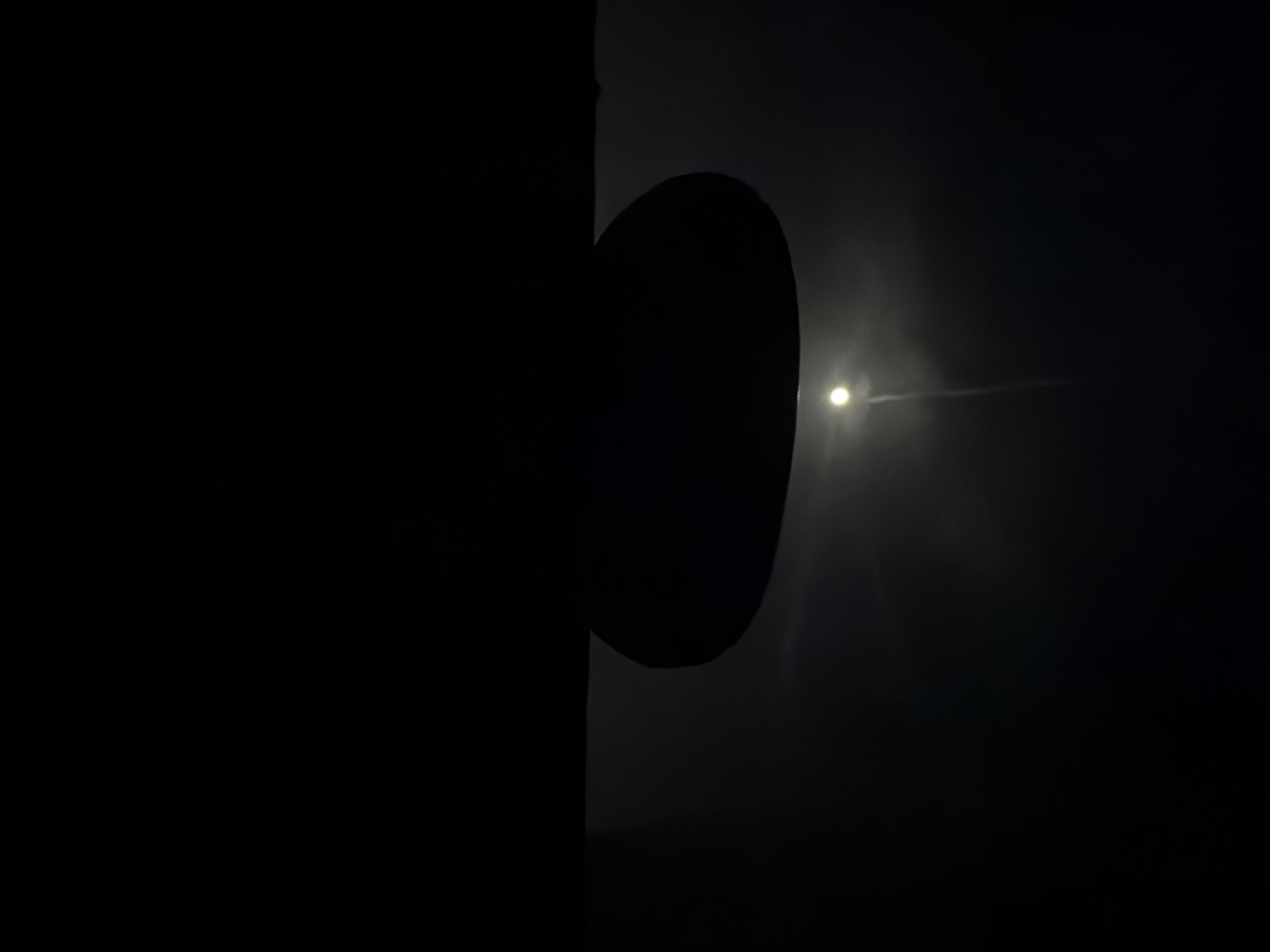 The image, likely taken at night in Sant Martí Sarroca, Spain, is dominated by shadows. A dark, opaque shape, seemingly attached to a vertical surface, occupies the middle of the frame. To the right, a point of light pierces the darkness, emitting a faint beam. The rest of the scene is enveloped in deep shadow.