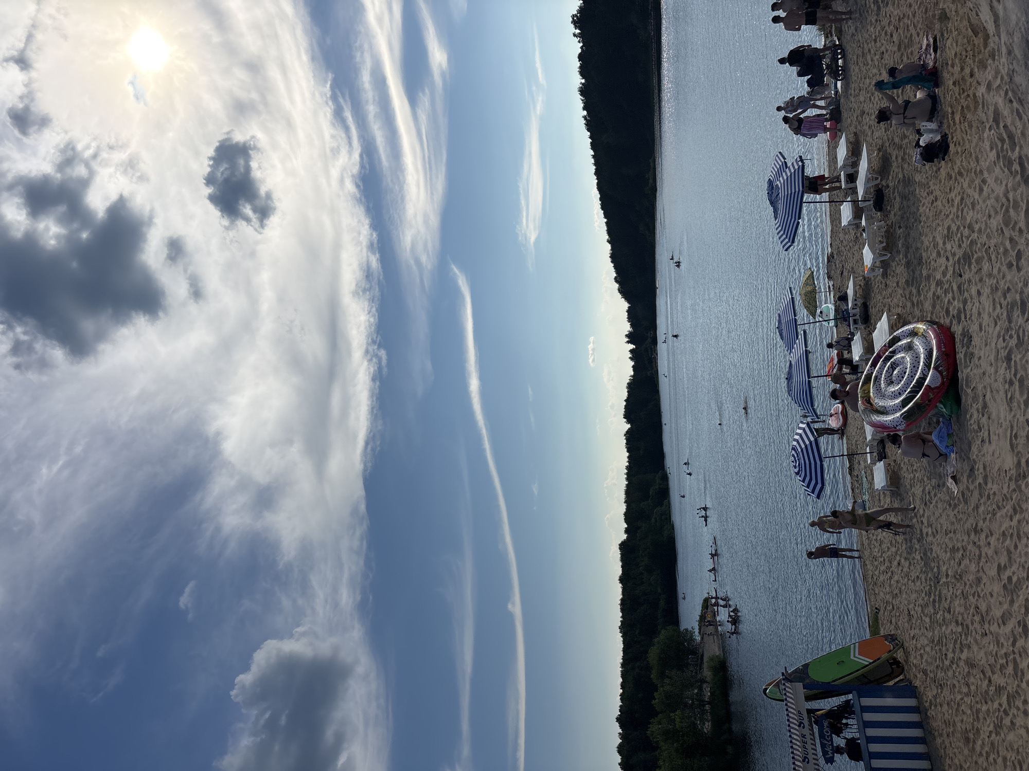 The image depicts a sunny day at a beach on a lake. The sky is a vibrant blue with scattered white and dark clouds, and the sun shines brightly. The beach is sandy and lined with trees, and the lake is calm with several people engaged in water activities like kayaking and paddleboarding.
On the sandy shore, beachgoers are relaxing on lounge chairs under striped umbrellas. Some individuals are standing or walking, and there are a few inflatable toys, including a large, circular one with a black and white spiral pattern. A few people are visible near a small, striped structure, possibly a beach concession stand.
The image appears to be taken in the afternoon, given the angle of the sun and the shadows cast. The atmosphere seems relaxed and leisurely, with people enjoying a day by the water. The location is identified as Kurovskoye, Russia. Visible text includes "SUPER SUP" and "Welcome" on a blue and white striped sign.