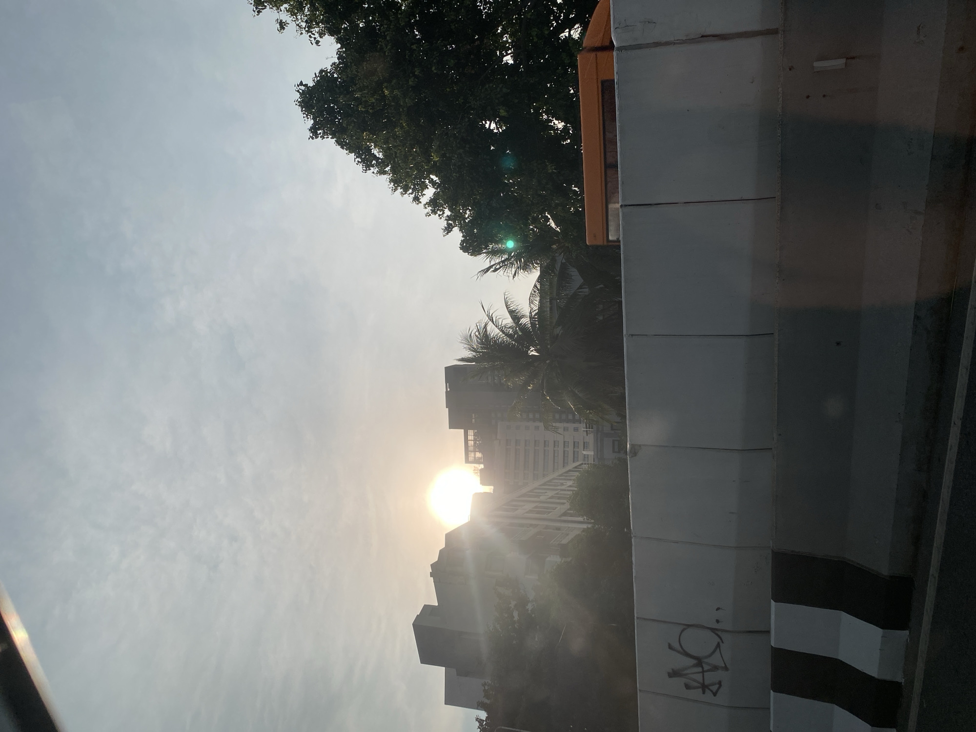 The image appears to be taken from a vehicle traveling along a road in Bangkok, Thailand. The sun is low in the sky, possibly during sunrise or sunset, positioned behind buildings. A concrete barrier is partially visible on the right, showing some graffiti. Trees are also visible, obscuring some of the buildings. The sky is partly cloudy.