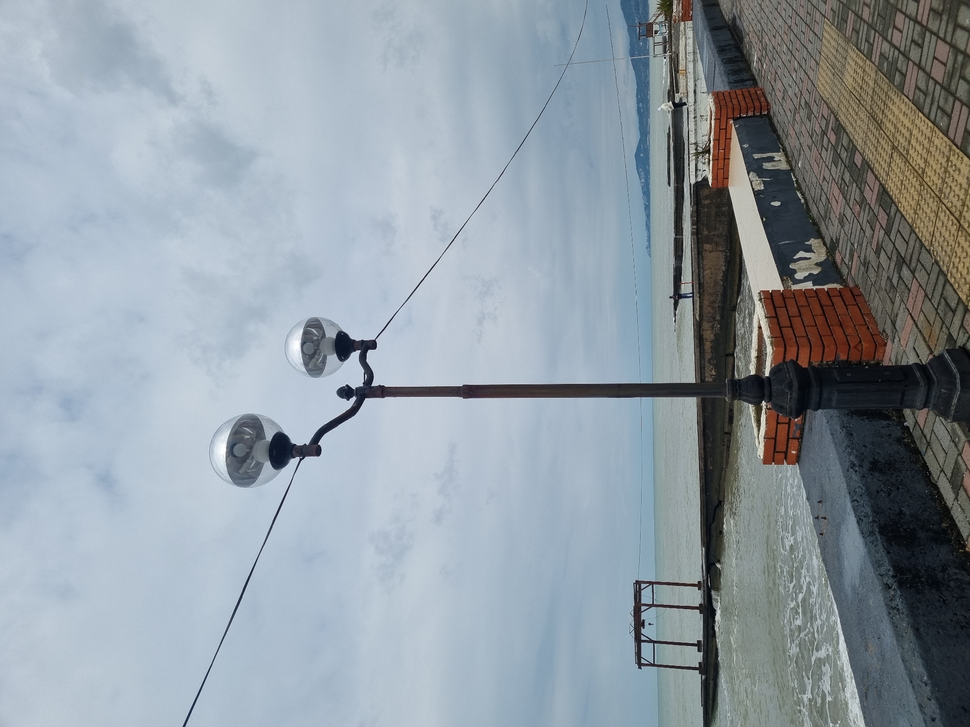 This image depicts a tranquil coastal promenade scene, likely located in Sochi, Russia, under an overcast daytime sky. The perspective looks along the coastline and out over a body of water.
In the right foreground, a tall street lamp stands prominently. It has a dark, possibly metal pole with two elegant, curved arms, each supporting a spherical, clear glass globe enclosing a light bulb. A thin, dark cable stretches diagonally across the sky from near the top of the lamp.
To the left of the street lamp, a paved promenade extends into the distance. The paving features a pattern of reddish-brown and gray bricks, with a notable strip of yellow tactile paving visible closer to the viewer. Adjacent to the promenade, bordering the water, is a low barrier or wall. This structure is composed of sections of dark-painted concrete, showing some signs of peeling white paint, interspersed with short columns constructed from reddish-orange bricks.
Beyond the promenade and the low wall, a calm, grayish-blue sea stretches towards the horizon. Gentle waves can be seen near the shore. In the far distance, across the water, a range of low-lying mountains or hills is faintly visible against the cloudy sky. Two small, indistinct figures appear to be walking near the water's edge further down the promenade.
Out in the water, a rust-colored, open-frame structure, possibly a small, disused pier or gantry, stands between the viewer and the distant mountains. The sky above is entirely covered with soft, light gray clouds, indicating an overcast but bright day. The overall mood is peaceful and calm. No visible text is present in the image.