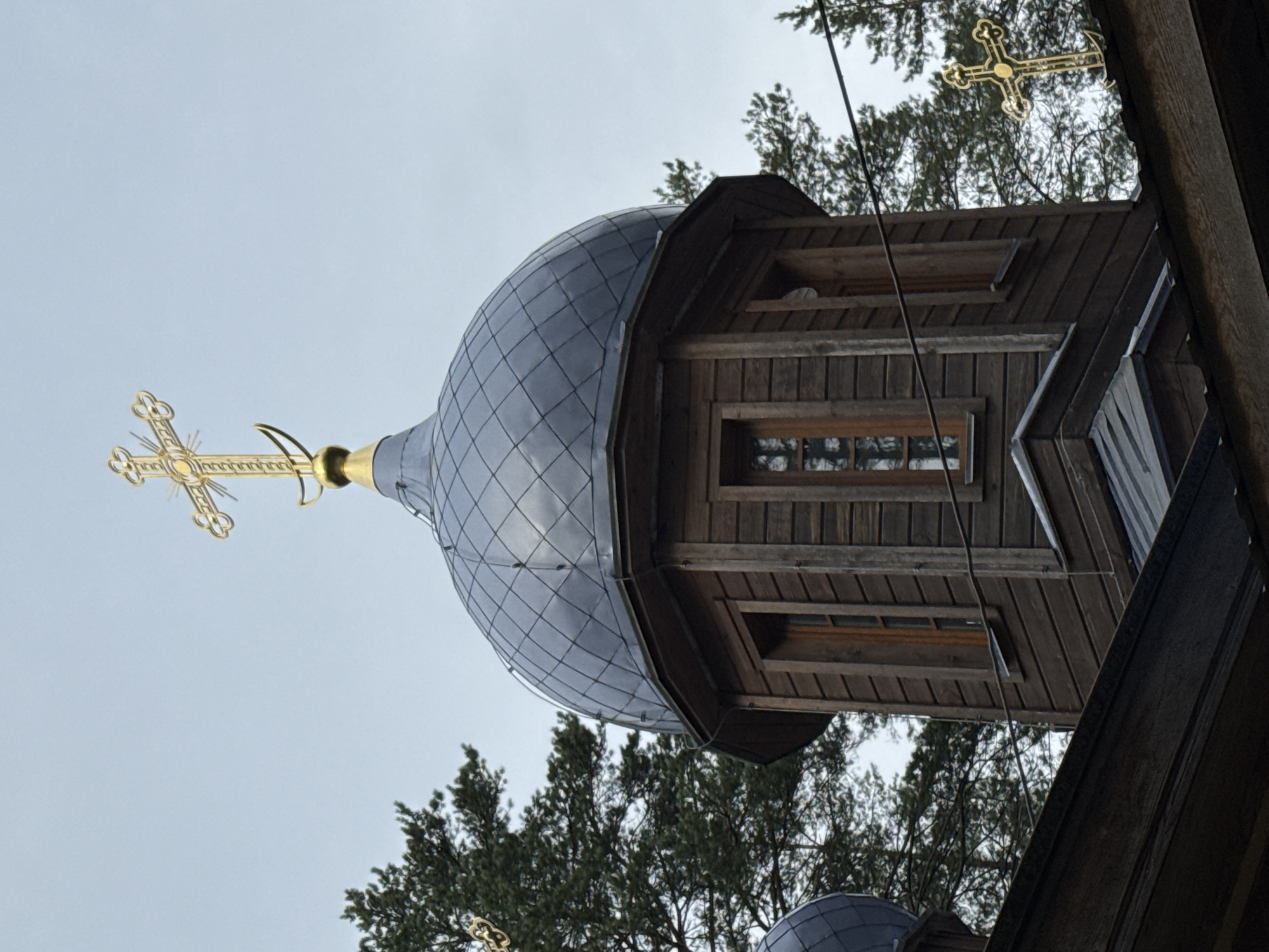 A close-up, low-angle shot shows the upper portion of a wooden Orthodox church in Nenimäki, Russia. The main subject is a dark gray, multi-faceted dome topped with a golden, ornate cross. The dome is supported by a hexagonal wooden tower with vertical wooden planks and narrow windows. In the background, dark green pine trees fill the sky, and a faint wire runs horizontally across the frame. Another smaller golden cross is visible on the roofline to the right. The sky is overcast and a pale blue.