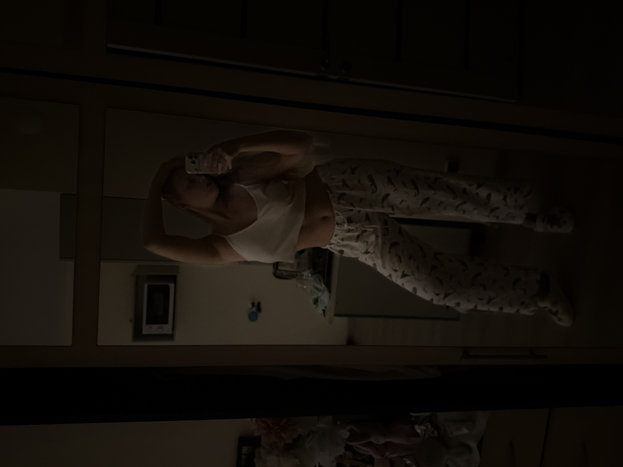 A reflection in a mirror shows a woman wearing a white tank top and patterned pajama pants, holding a smartphone in her hand, presumably taking a selfie. The environment is dimly lit, revealing sections of a room containing furniture such as a cabinet or dresser. There are other objects such as a picture frame on the wall, a bottle of liquid on a table, and a floral decoration on the floor. The location is unknown and cannot be determined from the image.