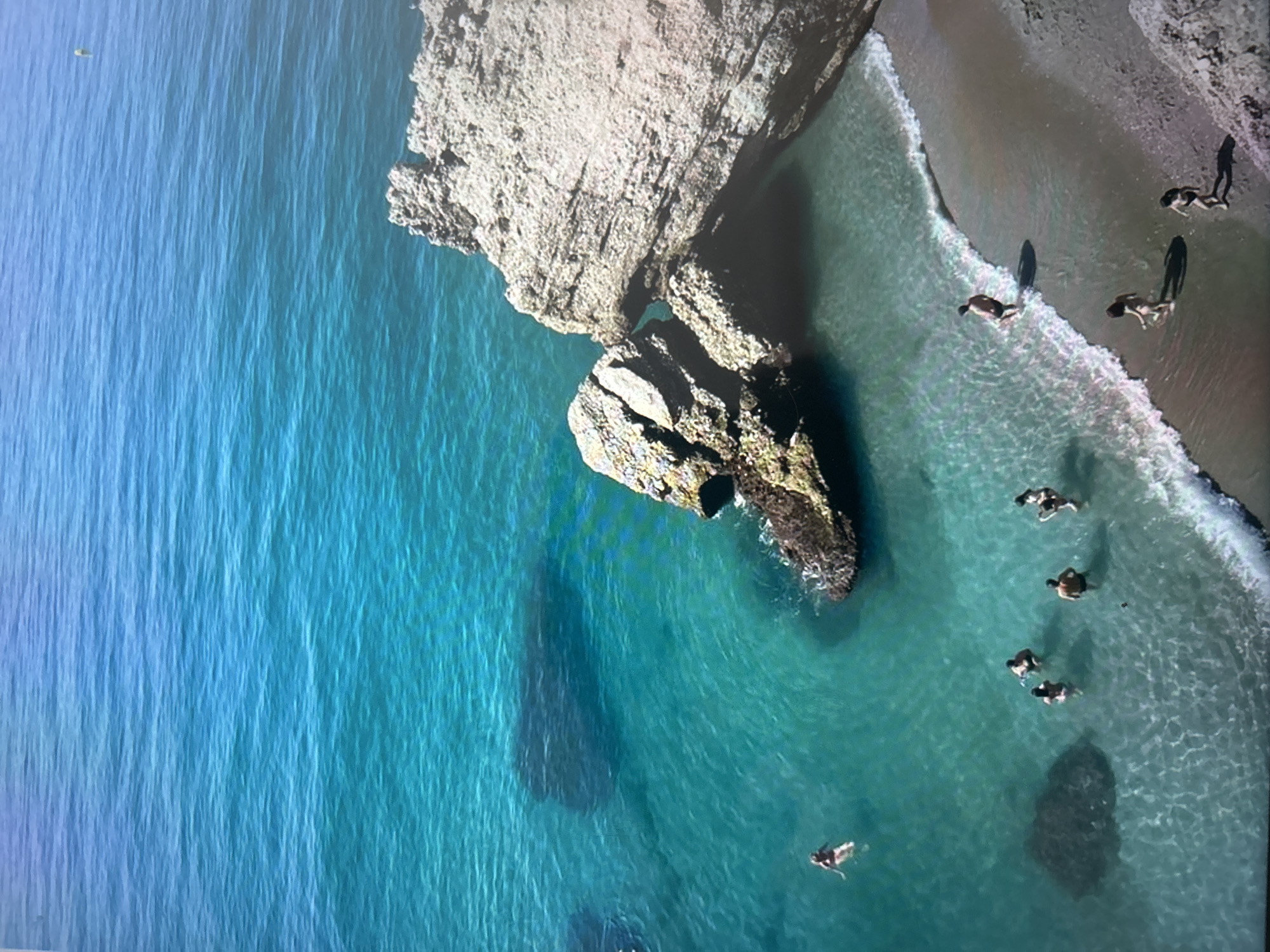 An aerial or high-angle photograph captures a vibrant coastal scene featuring clear, blue-green waters, rugged rocky formations, and a sandy beach. The image is oriented vertically, showing the ocean on the left and the coastline on the right.
The water transitions from a deep, rich blue in the deeper areas to a bright, clear turquoise and light green closer to the shore, indicating its pristine clarity. Gentle waves are seen breaking along the sandy edge. Several people are engaged in activities within and around the water. Some individuals are swimming or wading in the clear shallows, their forms visible beneath the surface. Others are on the sandy beach, with some appearing to walk or stand near the waterline, while one person appears to be lying down.
Prominent light-colored rock formations and cliffs extend into the water, casting significant dark shadows on the ocean surface, suggesting a relatively low sun angle, likely early morning or late afternoon. These shadows are also visible under the water, hinting at submerged rocks. The people on the beach also cast long, distinct shadows, reinforcing the inference about the time of day. The overall impression is one of a serene and natural beach environment, ideal for recreation and relaxation. No visible text is present in the image.
