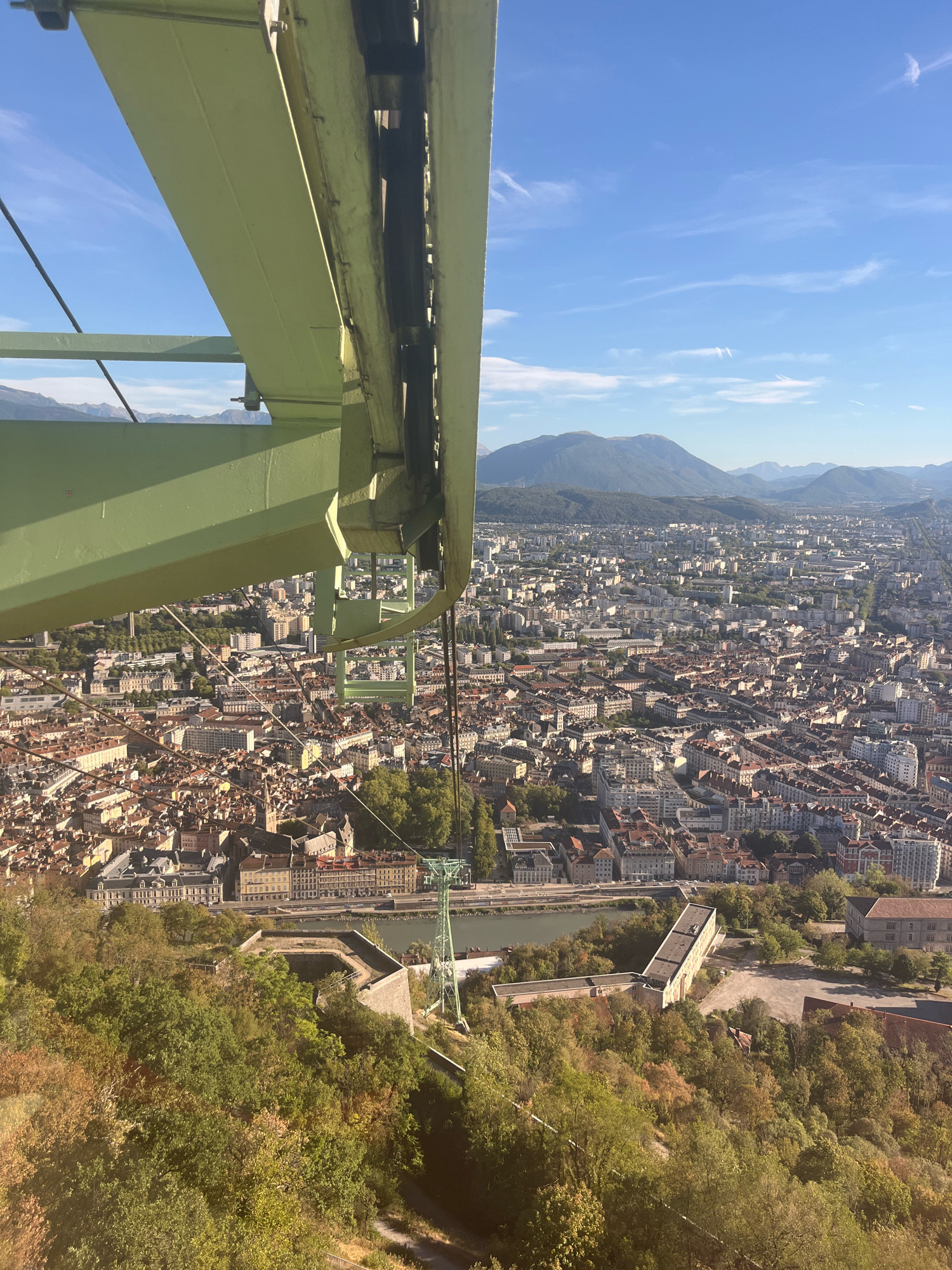 The image, taken from a cable car, offers a panoramic view of Grenoble, France, on a clear, sunny day. In the foreground, a portion of the green cable car structure is visible, with cables and mechanisms for its operation. Below, the cityscape of Grenoble unfolds, characterized by a dense arrangement of buildings with reddish-brown roofs. A river, likely the Isère, winds through the city, with bridges crossing it. The city is framed by rolling hills covered in lush green trees, and in the distance, a mountain range under a bright blue sky with wispy clouds. The perspective suggests the cable car is ascending or descending a hillside overlooking the city. No people are discernible in the image. The scene captures a scenic and urban environment from a high vantage point.