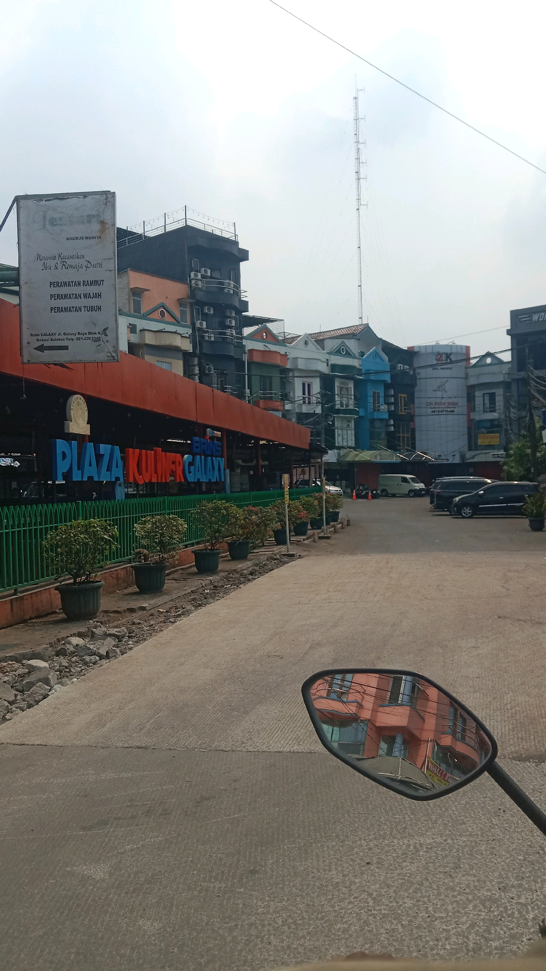 The image shows a street view in the city of Bekasi, Indonesia, specifically in the Galaxy area. In the foreground, a motorcycle's side mirror reflects an orange building with windows. The street itself is paved with concrete, showing some signs of wear and tear, with piles of debris and construction materials on the left side.
Dominating the midground is a row of buildings, some of which appear to be shops or restaurants. A prominent sign reads "PLAZA KULINER GALAXY" in large, colorful letters. In front of the plaza, a green metal fence encloses a line of potted plants. Behind the plaza, several multi-story buildings are visible, with varying architectural styles and colors, including a tall communication tower in the distance. The sky is overcast.
On the left side of the image, a white sign with black text provides information about beauty services, listing "PERAWATAN RAMBUT," "PERAWATAN WAJAH," and "PERAWATAN TUBUH" (hair, face, and body treatments). The sign also includes an address and phone number. Several cars are parked on the street. The overall impression is a bustling urban environment with commercial activity.