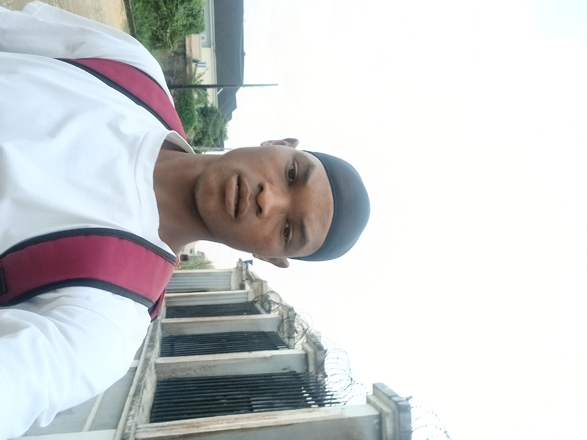 A medium shot captures a young Black man from the chest up, looking directly at the camera. He wears a black skullcap and a white t-shirt with a maroon backpack slung over his shoulders. The background features a light-colored, overcast sky, green trees, a house with a dark roof, and a fence with vertical bars and coiled barbed wire. The man's face is the primary focus, with his expression neutral. The overall scene suggests an outdoor setting in a residential area.