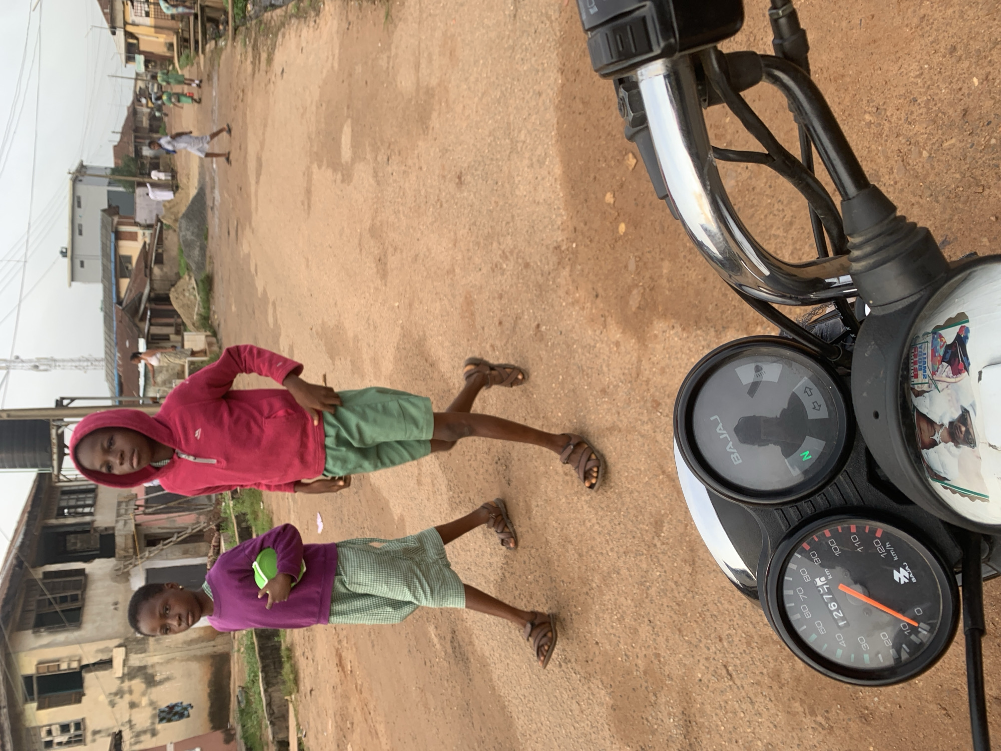In Shagamu, Nigeria, two children walk along a dirt road. The child on the left, wearing a purple long-sleeved shirt and a green and white checkered skirt, carries a green object. The child on the right, in a pink hooded sweatshirt and green shorts, walks ahead. The scene is framed by buildings and other people in the background. A motorcycle's handlebars and dashboard are in the foreground, with the speedometer showing 12675 km.