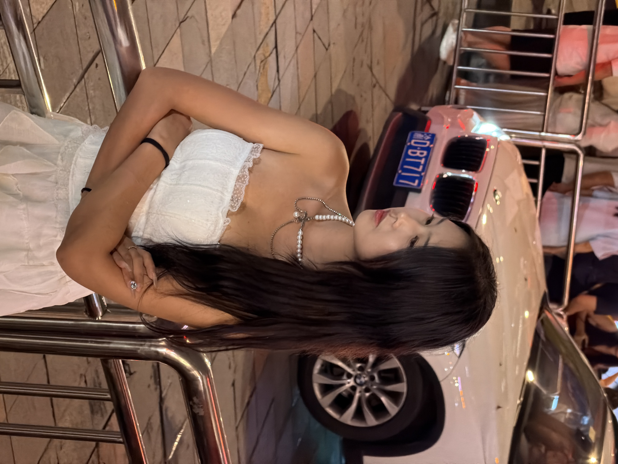 The image shows a young woman in a white dress. She is leaning against a metal railing, with her arms crossed. Her hair is long and dark, and she is wearing a pearl necklace. A white car is parked next to her, with its license plate visible. The license plate reads "D-B717". The scene appears to be outdoors at night, with streetlights reflecting on the car and the railing. The woman has a serene expression on her face, and her posture suggests a moment of quiet contemplation. The background is blurred, but it appears to be a city street. The overall mood of the image is one of relaxed elegance. The setting is likely Yuzhong District, China, as indicated by the provided location context.