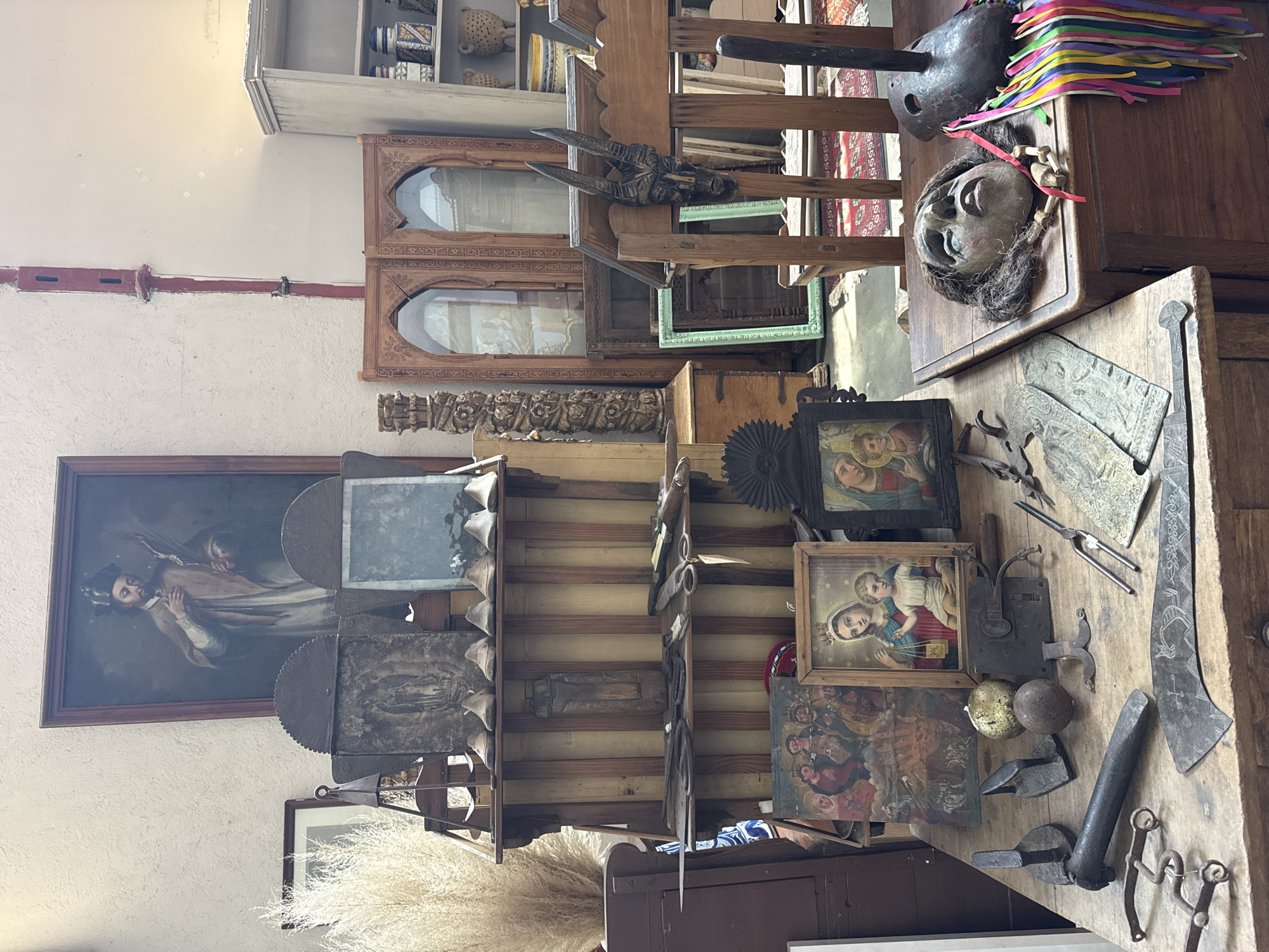 The image shows a cluttered interior, likely a store or storage area, filled with antique and vintage items. The location is suggested to be San Miguel de Allende, Mexico, but this cannot be confirmed from the visual details alone.
A painting depicting a man with a crown hangs on the wall. In front of it is a wooden shelf unit holding various objects. Near the top right, there is a wooden table covered with more items, including framed religious paintings, metal tools, and decorative objects. There is a mask with hair on the table and a colorful decoration behind it.
The overall environment suggests an eclectic collection of artifacts and decorative pieces.