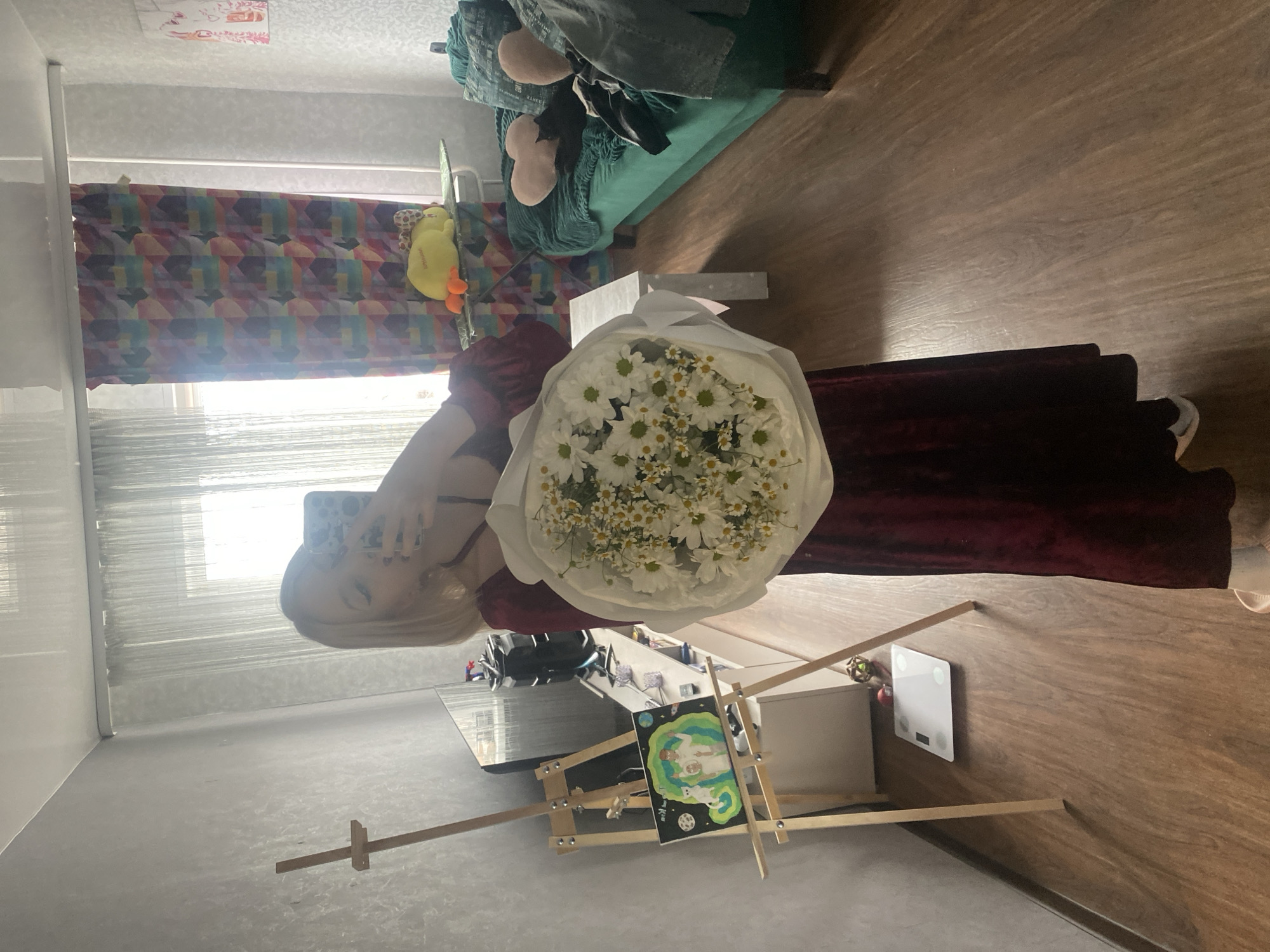 A woman with long blonde hair is holding a bouquet of white daisies wrapped in white paper in an indoor setting. She is wearing a dark red garment. An easel with a painting stands near a white console table and a white digital scale on a brown wooden floor. In the background, there is a patterned window curtain and a green couch with stuffed animals. The image appears to be taken in a residential location, potentially in Izhevsk, Russia. The location is specified because the user has requested it in the prompt.