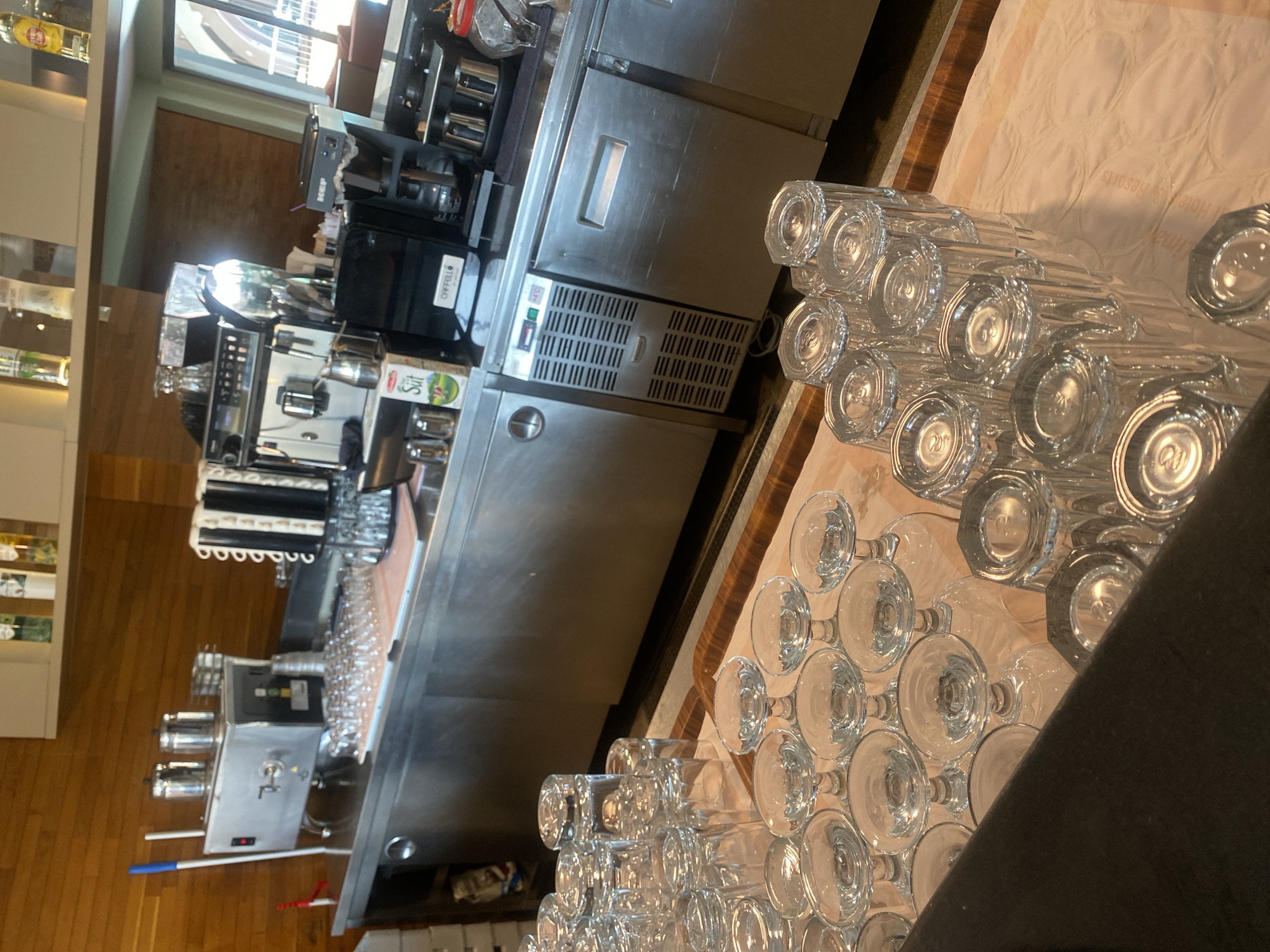 The image shows a bar or restaurant counter with various items. On the counter, there is a coffee machine, a beverage dispenser, and multiple stacks of clean glassware, including wine glasses and tumblers. The background features wooden paneling and shelves stocked with bottles. The overall scene suggests a commercial establishment, possibly in Aksu, Turkey, preparing for service or recently cleaned.