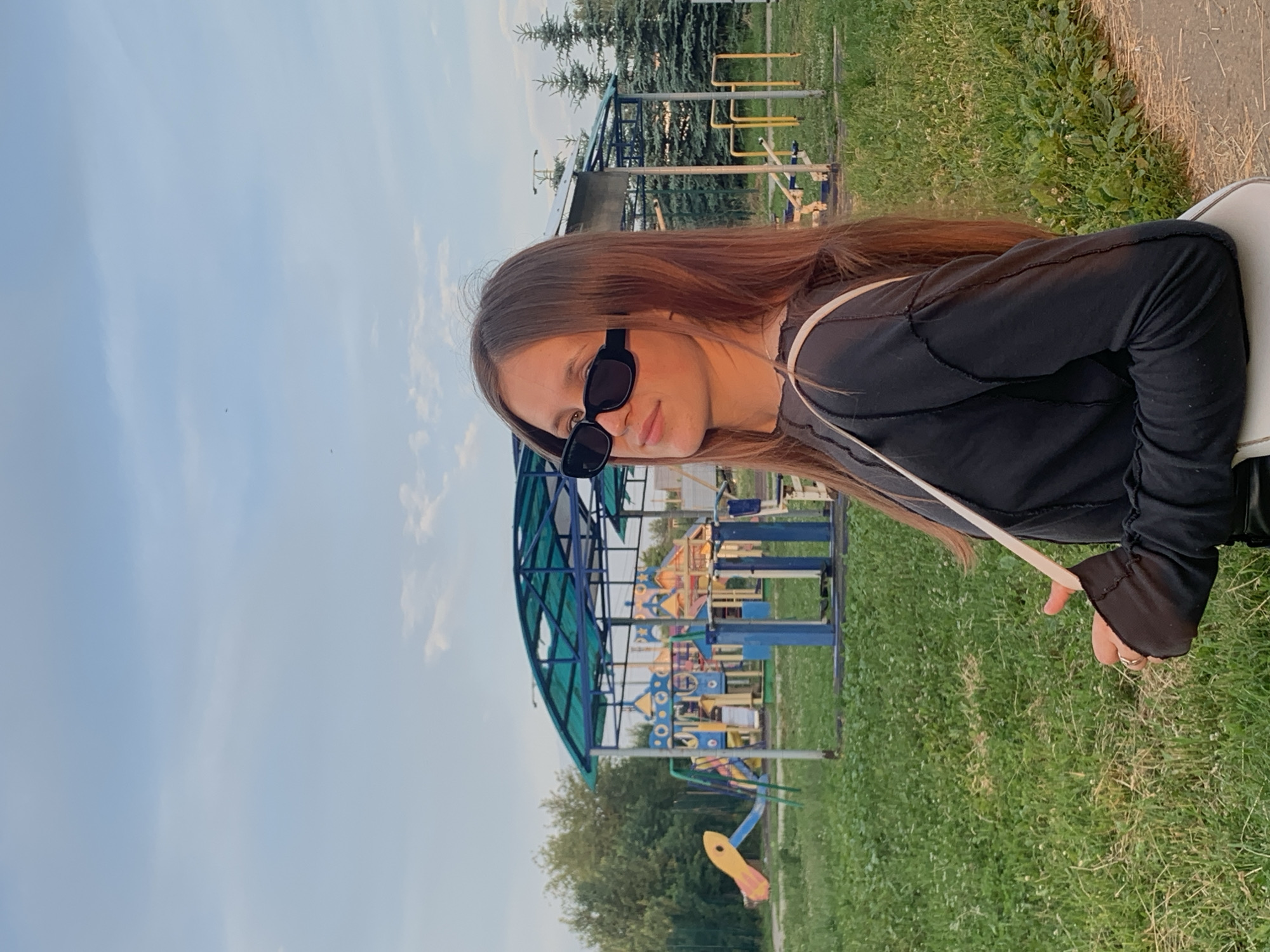 A young woman with long, brown hair and black sunglasses is standing in a park with a playground in the background. She is wearing a black long-sleeved shirt and a white purse. The playground has various colorful structures, including slides and swings. The scene appears to be in the city of Fokino, Russia, based on the provided information. The lighting suggests it is late afternoon or early evening.