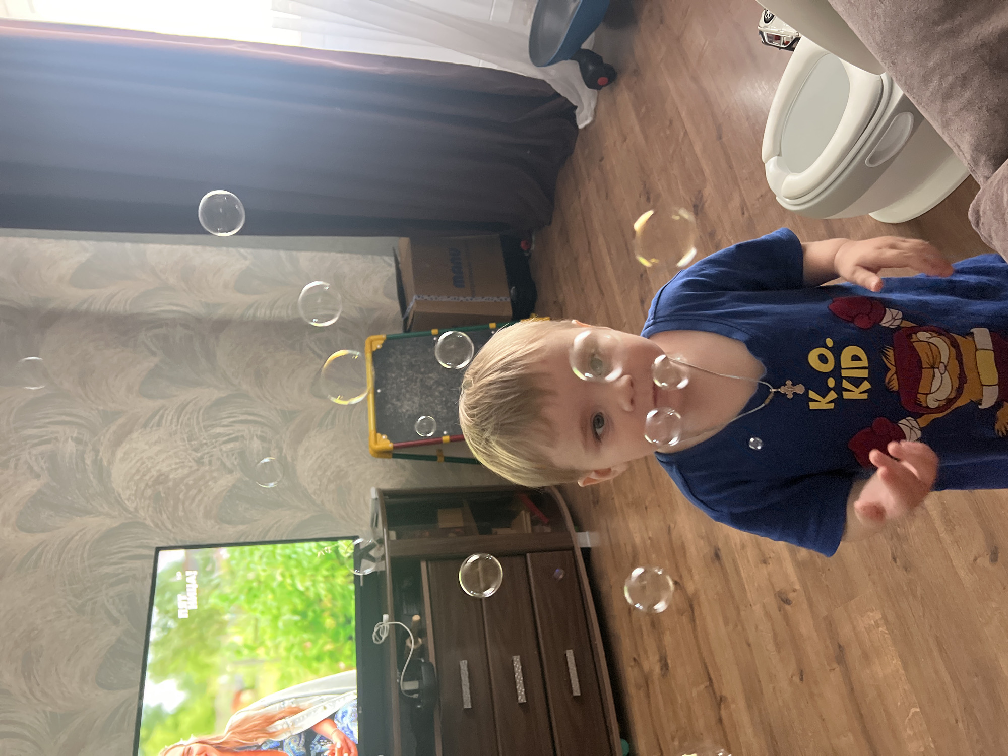 The image captures a young boy, appearing to be approximately two to three years old, in a living room setting. He has blonde hair and is wearing a blue t-shirt with a cartoon character graphic and the text "K.O. KID". The boy is looking towards the camera with an open mouth, seemingly interacting with soap bubbles that are floating around him. His right hand is extended forward.
The environment includes a wooden floor, a dark wooden entertainment center with drawers to the left, and a television screen displaying a program with a woman. To the right of the boy, a white and grey potty training seat is visible. Further to the right, a dark curtain hangs near a window with sheer white curtains. In the background, there is a cardboard box with writing on it and a child's easel. The room's wallpaper has a textured, abstract pattern.
The image was taken indoors, likely in a residential space within the city of Noviy, Russia. The lighting suggests it is daytime, with natural light coming from the right side of the frame. The focus is on the boy and the floating bubbles, creating a sense of playful activity.