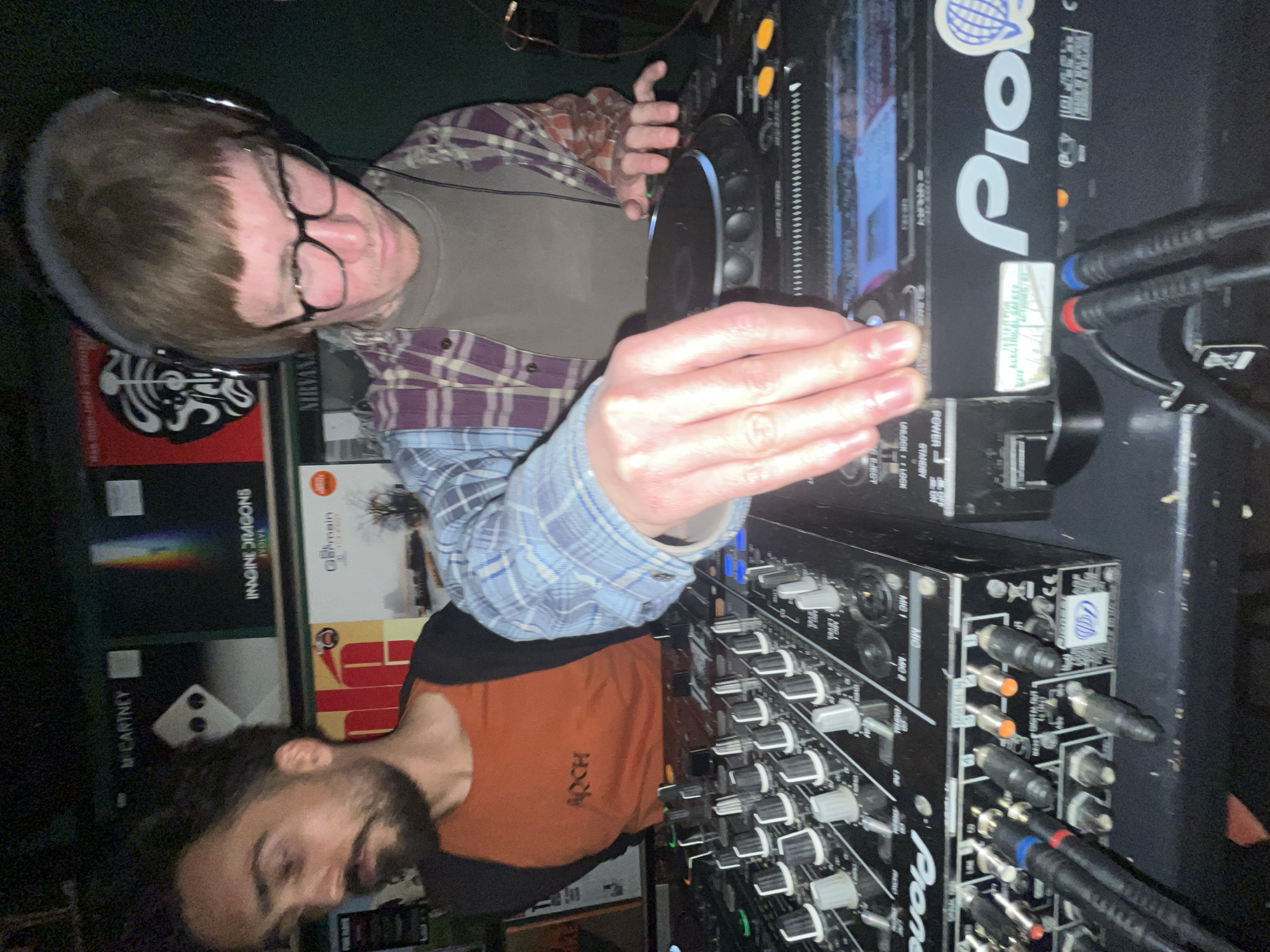 The image depicts two people in an indoor setting, possibly a club or a music venue. One person, wearing glasses and headphones, is standing near a Pioneer DJ setup, manipulating the controls. He is wearing a plaid shirt. The second person, with a beard, is wearing an orange and black shirt with "ARCH" printed on it. There are vinyl records visible in the background, including albums by Nirvana, Pink Floyd, Imagine Dragons, and Paul McCartney. The DJ equipment includes a mixer and a CDJ player, with various cables connected. The exact location within Budva, Montenegro, cannot be determined from the image.