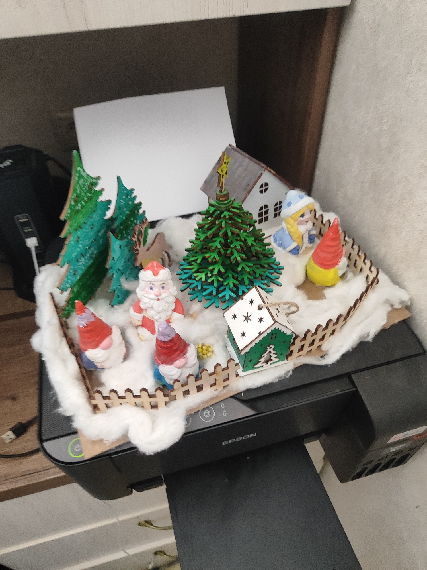 An overhead shot showcases a Christmas-themed diorama placed atop a black EPSON printer. The diorama features a winter scene with fluffy white material simulating snow, scattered around miniature wooden Christmas trees, festive figures including Santa Claus and two gnomes, a young girl in a blue hat and coat, and a small green house with star cutouts. A wooden fence encloses the scene. The background reveals a light-colored wall and a portion of a wooden shelf or cabinet. The printer has visible buttons and ports, and a black cable with a USB connector is plugged into it.