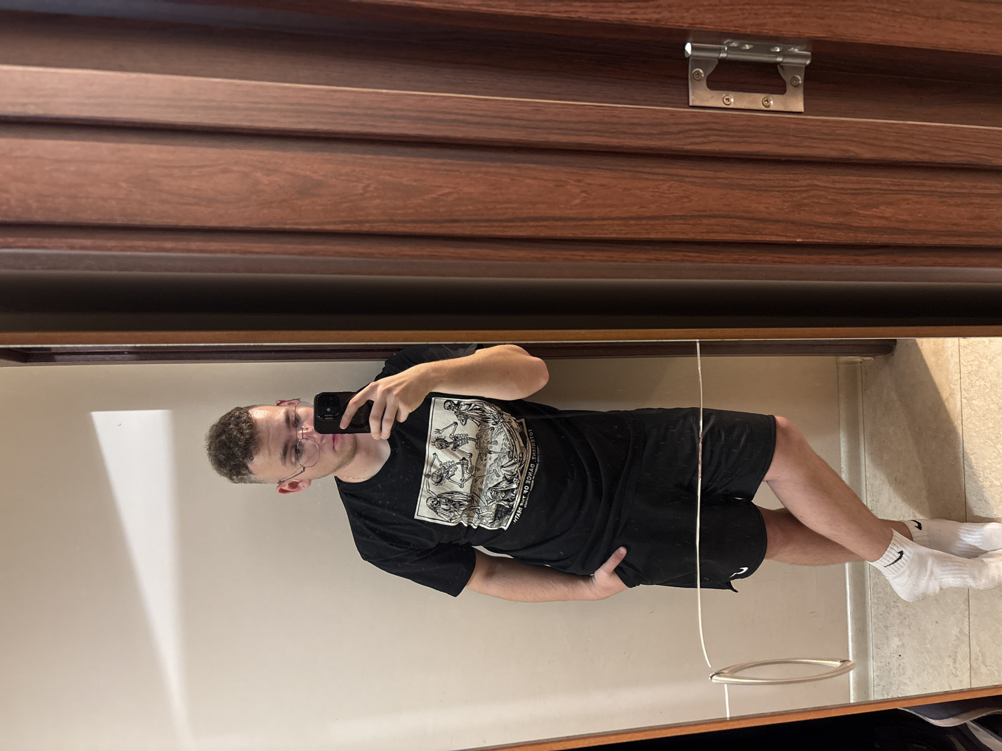 A man with short curly hair and glasses is standing in front of a mirror, taking a selfie with his phone. He is wearing a black t-shirt with a graphic print, black shorts, and white socks with a black Nike logo. The mirror is framed in dark wood, and the background shows a plain wall. Above the mirror is a dark wood cabinet with silver hardware. The location appears to be indoors, but the city and country cannot be determined from the image.