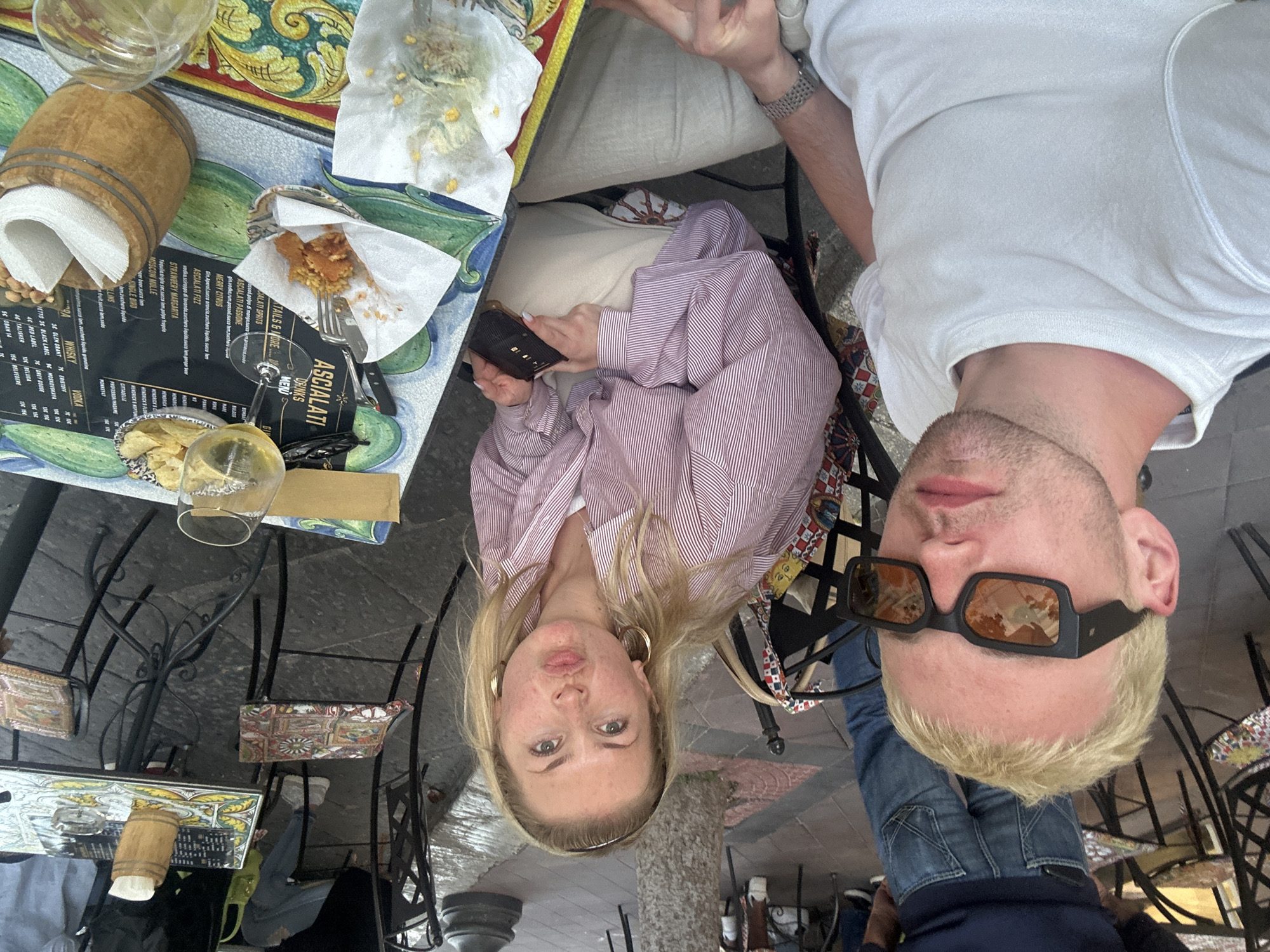 The image captures two young adults seated at an outdoor cafe table in Lipari, Italy, enjoying a casual meal or drinks. A woman with blonde hair, wearing a red and white striped long-sleeved shirt, is seated on the left, holding a smartphone and looking towards the camera with a slight pout. Opposite her, a man with short blonde hair and a white t-shirt wears dark rectangular sunglasses, also looking towards the camera.
The table features a colorful, tiled or painted pattern with green and yellow designs. On it are various items: a wooden barrel-shaped napkin holder, two wine glasses (one containing a pale liquid, likely white wine, next to a small bowl of potato chips), and plates with partially consumed fried snacks served on paper. Crumbs suggest food has been eaten. A black menu, prominently titled "ASCIALATI DRINKS MENU," is open on the table.
The menu lists a range of cocktails and spirits, including "ASCIALATI SPRITS," "ASCIALATI PASSIONE," "MERRY CITRUS," "ASCIALATI FIZZ," "STRAWBERY MARGARITA," and "MOSCOW MULLE." It also details various whisky and vodka brands such as GLEN GRANT, BLACK LABEL, RED LABEL, TALISKER, ERISTOFF, MOSKOVSKAYA, GREY GOOSE, BELUGA, and BELVEDERE, with prices next to them.
The chairs have ornate metal frames and decorative, patterned fabric seats. The ground below appears to be dark paving. The scene depicts a relaxed daytime gathering in an outdoor setting.