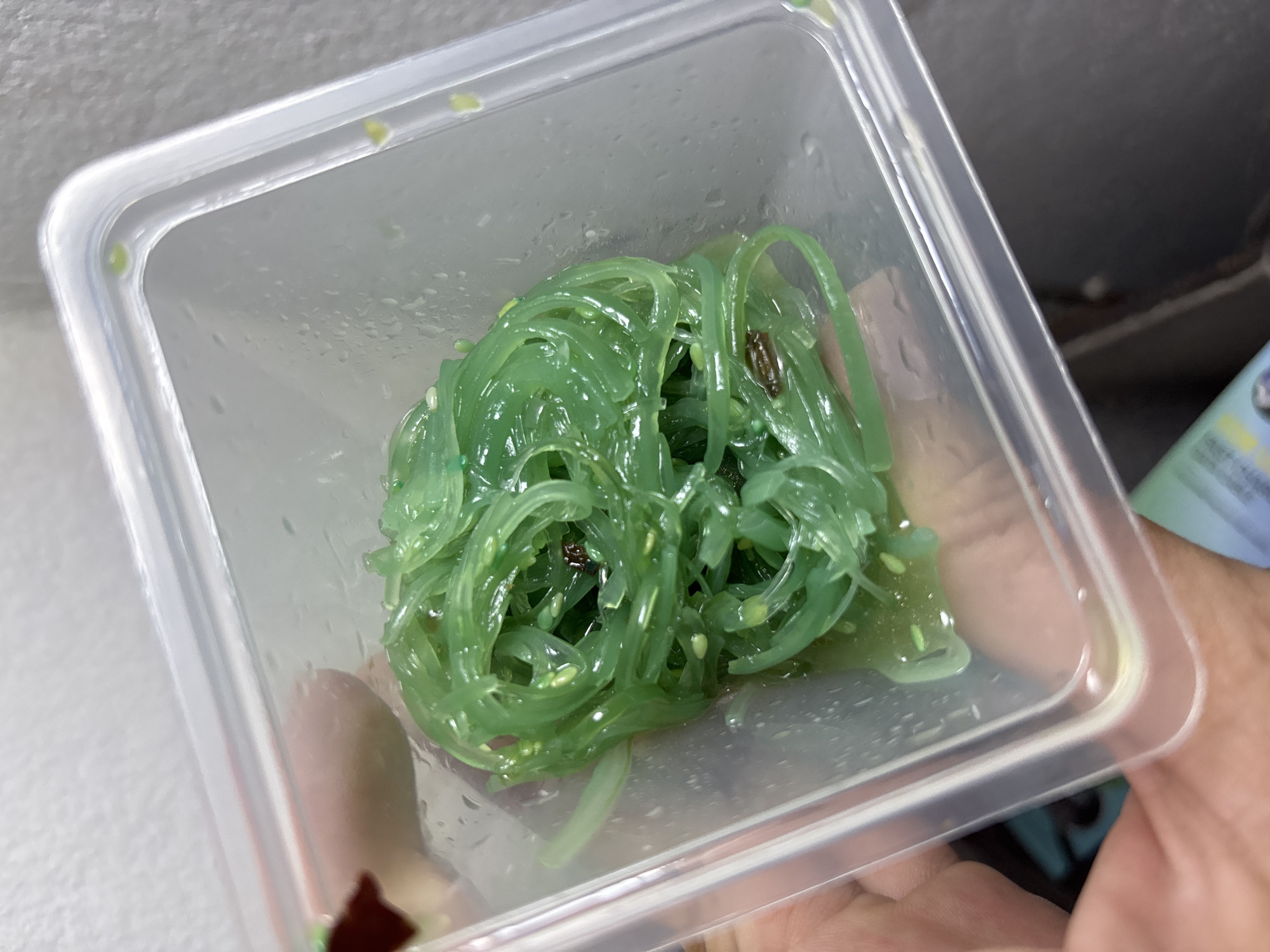 The image shows a close-up of a clear, square plastic container filled with green seaweed salad. The salad appears to be shiny and has sesame seeds mixed throughout. There's condensation on the inside of the container. Part of a hand is visible holding the container, and portions of a white surface and another object are visible in the background. The location is บ้านบางรักษ์, Thailand.