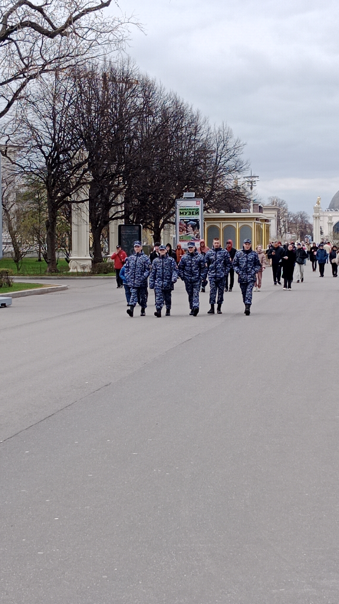 The image depicts a group of five individuals walking in a straight line on a wide paved road. They are wearing matching dark blue patterned uniforms and caps. Other people are walking in the background in the same direction, some in smaller groups and some individually. In the background, there are trees with bare branches, buildings with white columns, a gold-colored statue atop a building, a kiosk, and a sign that reads "МУЗЕЙ" (Museum) in Russian. The road stretches into the distance under a cloudy sky. The scene appears to be in a public park or square in Moscow, Russia.
