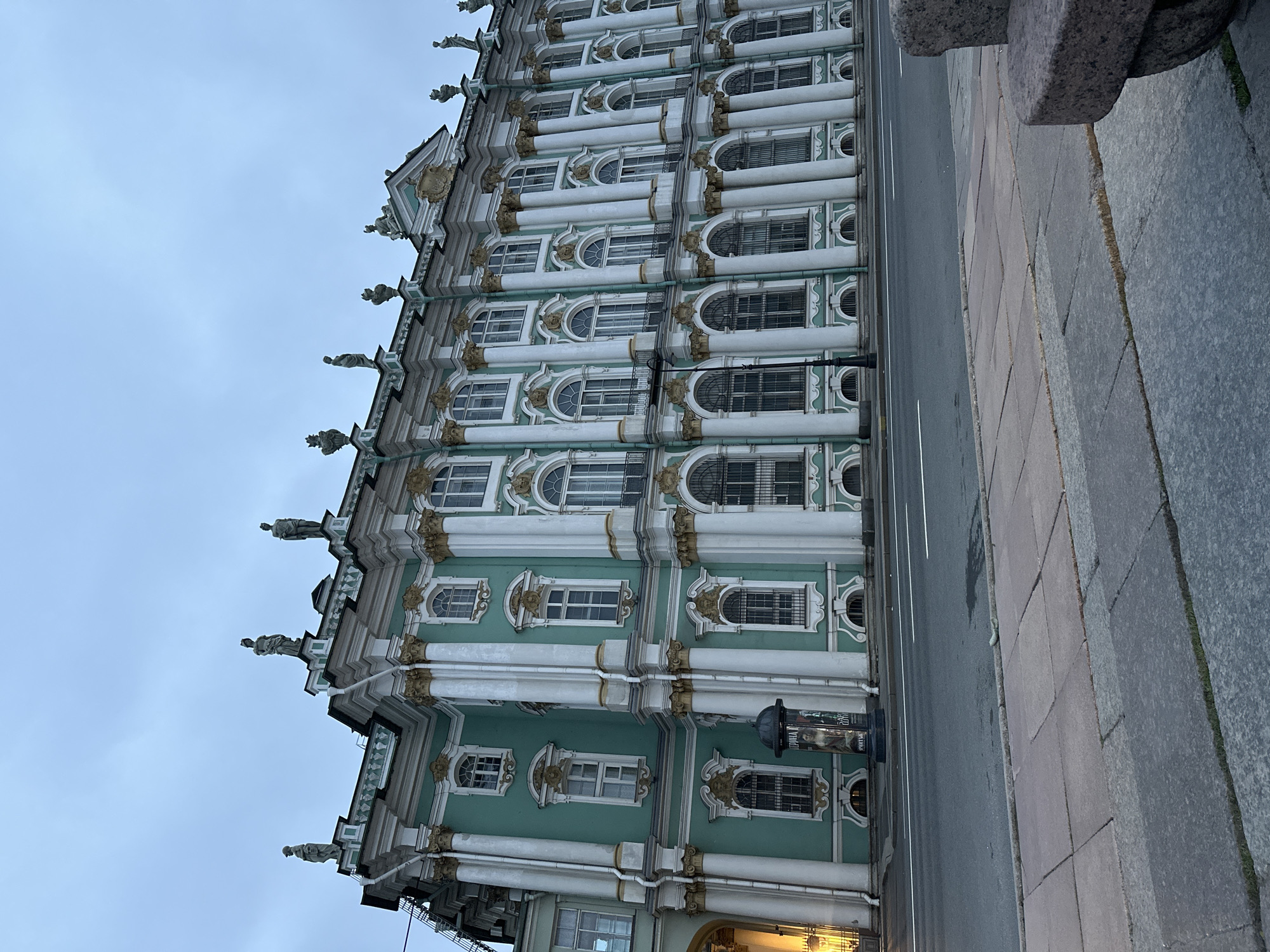 The image shows a detailed view of a large, ornate building with a turquoise facade and white columns. The building has multiple stories with numerous windows, many of which are arched. Sculptures of figures adorn the roofline. In front of the building is a street with a sidewalk and a paved area. A lamppost stands on the sidewalk, and a cylindrical advertisement pillar is visible near the building's entrance. The sky is overcast.