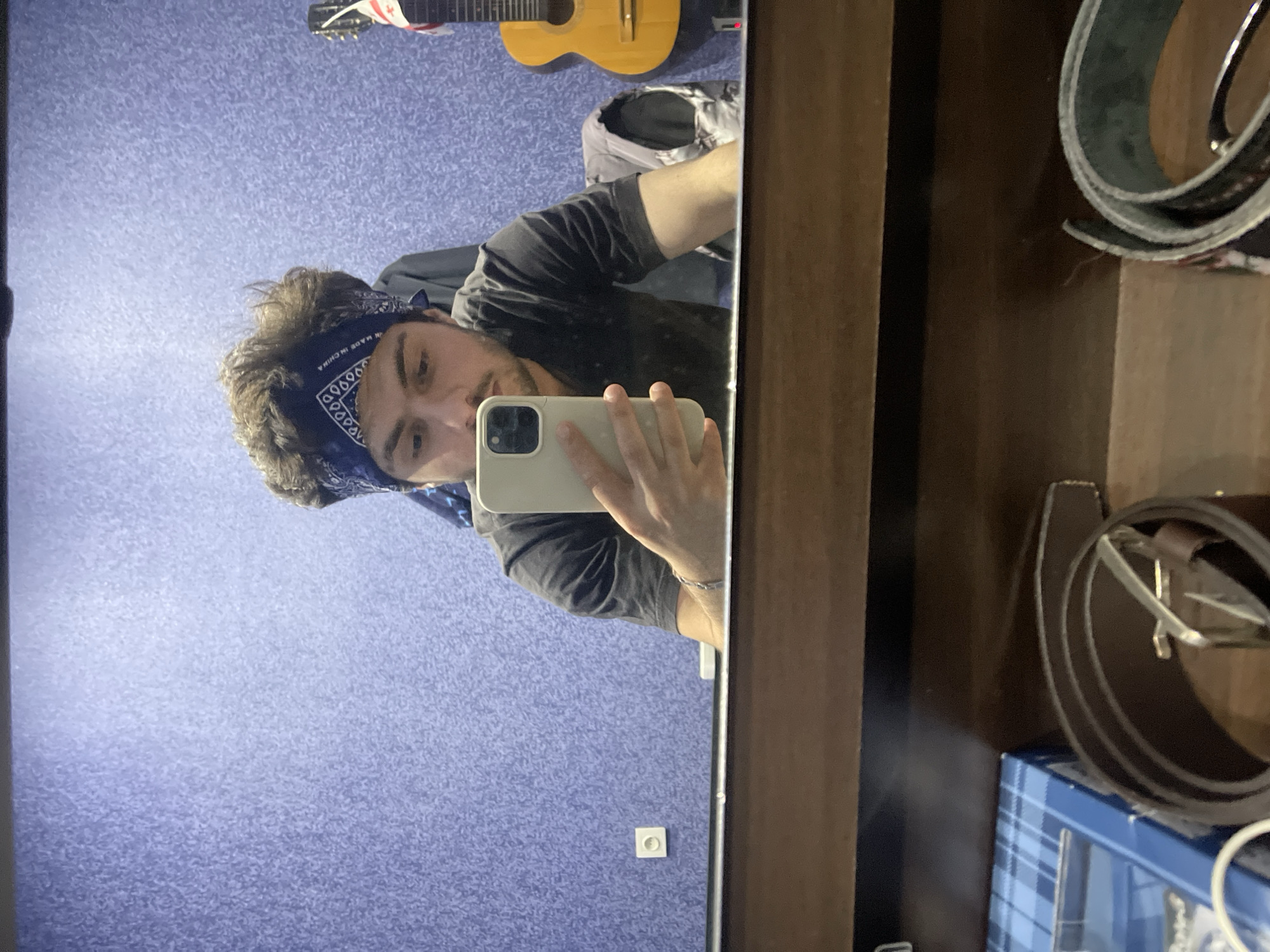 The image, taken from a mirror's reflection, shows a young man with dark, curly hair wearing a blue bandana and a dark t-shirt. He is holding a white smartphone in his right hand, appearing to take a selfie. His left arm is visible, with a silver bracelet on his wrist. In the background, a textured purple wall is visible, with a white electrical outlet on the left side. To the right, the neck and body of a classical guitar with a flag sticker (likely the Georgian flag) are partially visible. The foreground of the reflection reveals a wooden surface with various items, including a blue checkered box and two rolled-up belts. Based on the visible Georgian flag on the guitar, the image was likely taken in Tbilisi, Georgia.