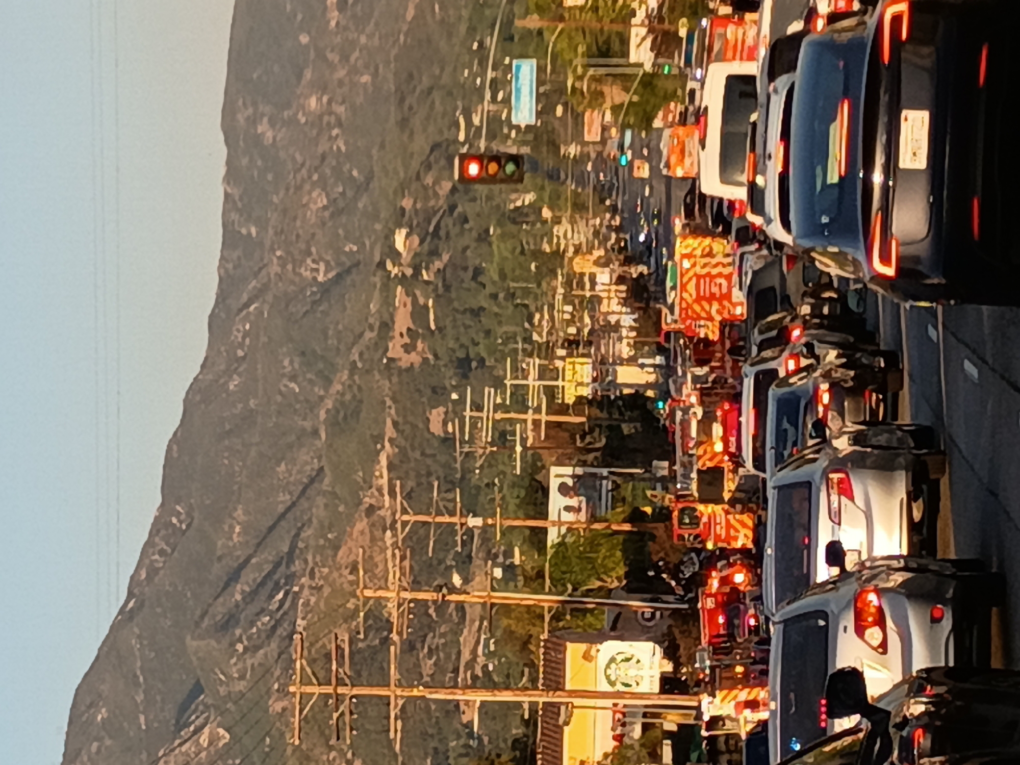 The image depicts heavy traffic congestion on a street in Los Angeles, California. A mountain range is visible in the background to the left. A Starbucks is situated along the street. Several emergency vehicles, including fire trucks and ambulances, are present, suggesting a potential incident or accident. Traffic signals are visible, one of which is red. Utility poles line the street.
