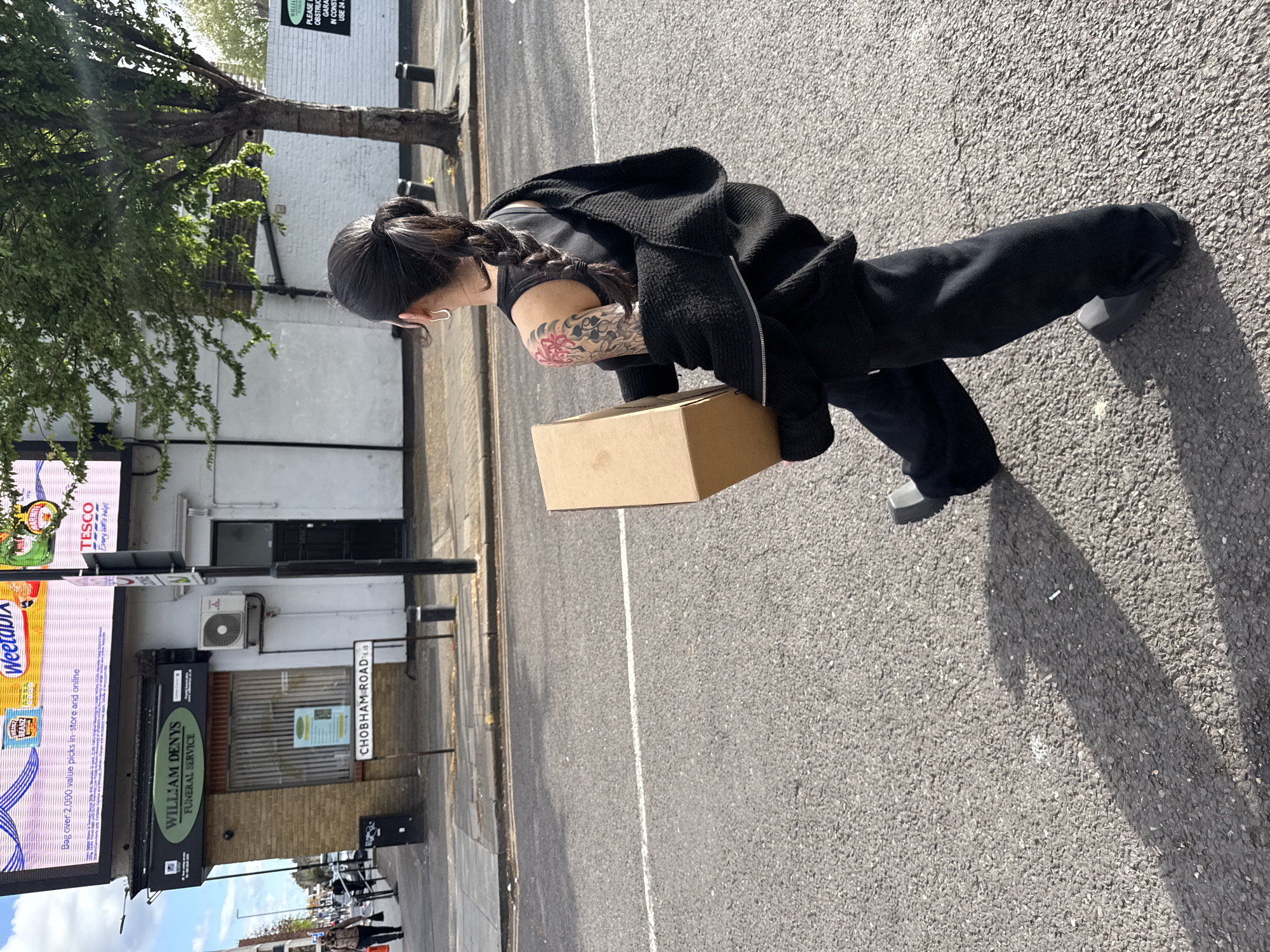 A woman stands on the side of Chobham Road in London, carrying a cardboard box. She has dark hair tied in a braid, a black jacket, and tattoos on her arm. She stands in front of the William Denys Funeral Service, next to a large electronic billboard advertising Tesco and Weetabix. The background includes trees and a building, and the street is paved.