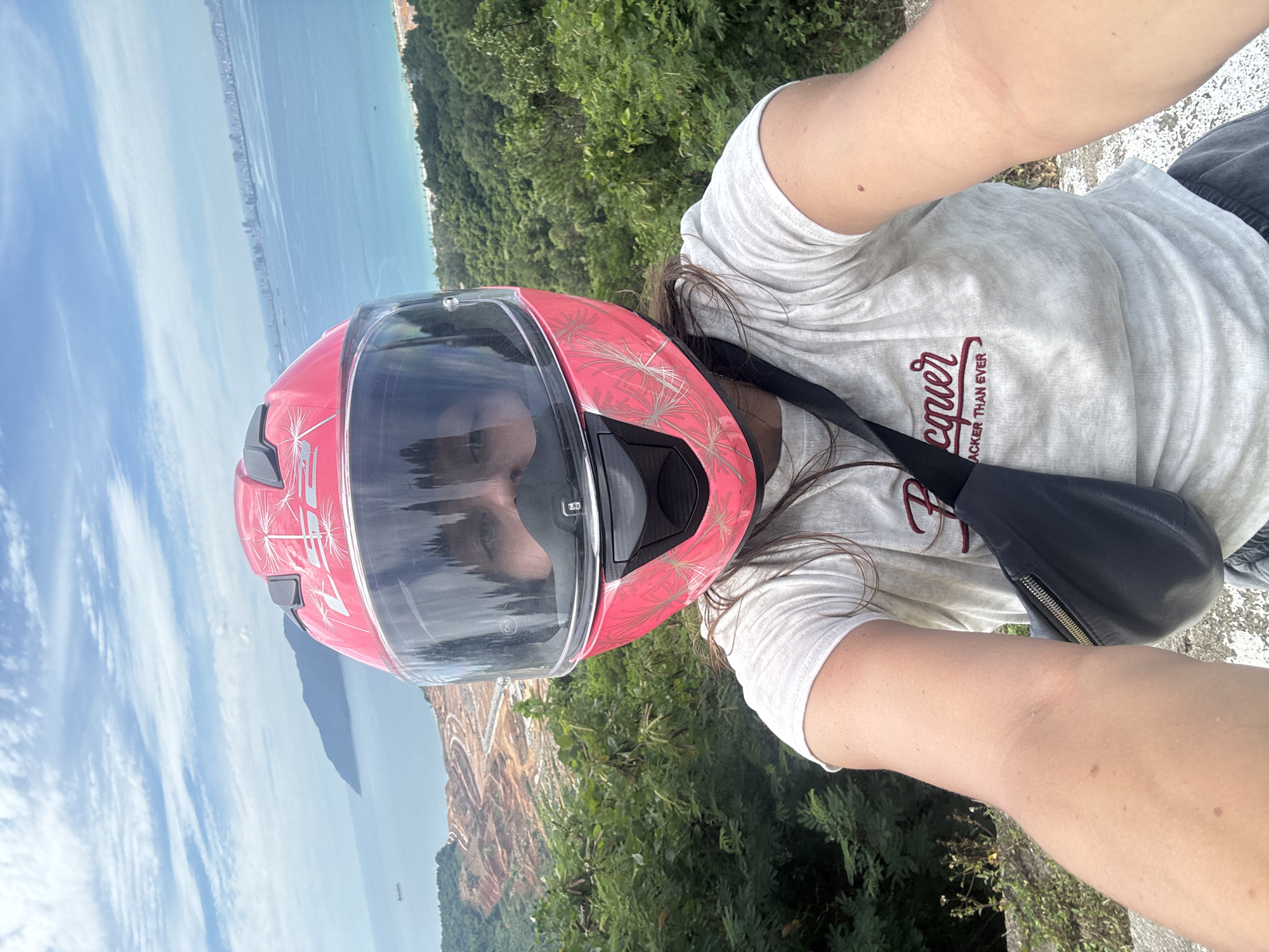 A woman is taking a selfie while wearing a pink motorcycle helmet and a white t-shirt. She is standing on a hill overlooking a coastal city in Đà Nẵng, Vietnam. In the background, there is a body of water with a few ships, a distant city skyline, and a construction site on the coast. The sky is partly cloudy. The woman has brown hair and is wearing a black crossbody bag. The t-shirt has red lettering that says "cquerACKER THAN EVER". The overall scene suggests a scenic view and a casual outing.