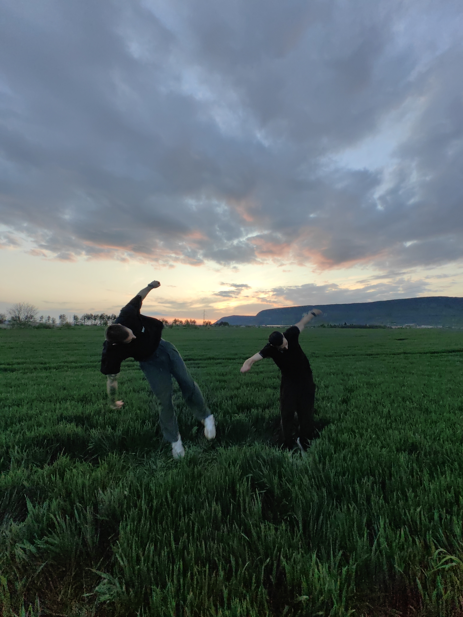 Two people are standing in a field of green grass. The person on the left is wearing a black t-shirt, grey jeans and white shoes, and is leaning to the left with their right arm raised and their left arm lowered. The person on the right is wearing all black, with a backwards black baseball cap and is bending to the right with their left arm raised and their right arm lowered. The background shows a sunset with pink, grey, and blue clouds. A few trees are to the left in the distance, and a mountain is visible in the distance to the right. The scene is located outside of the city of Shumen, Bulgaria.