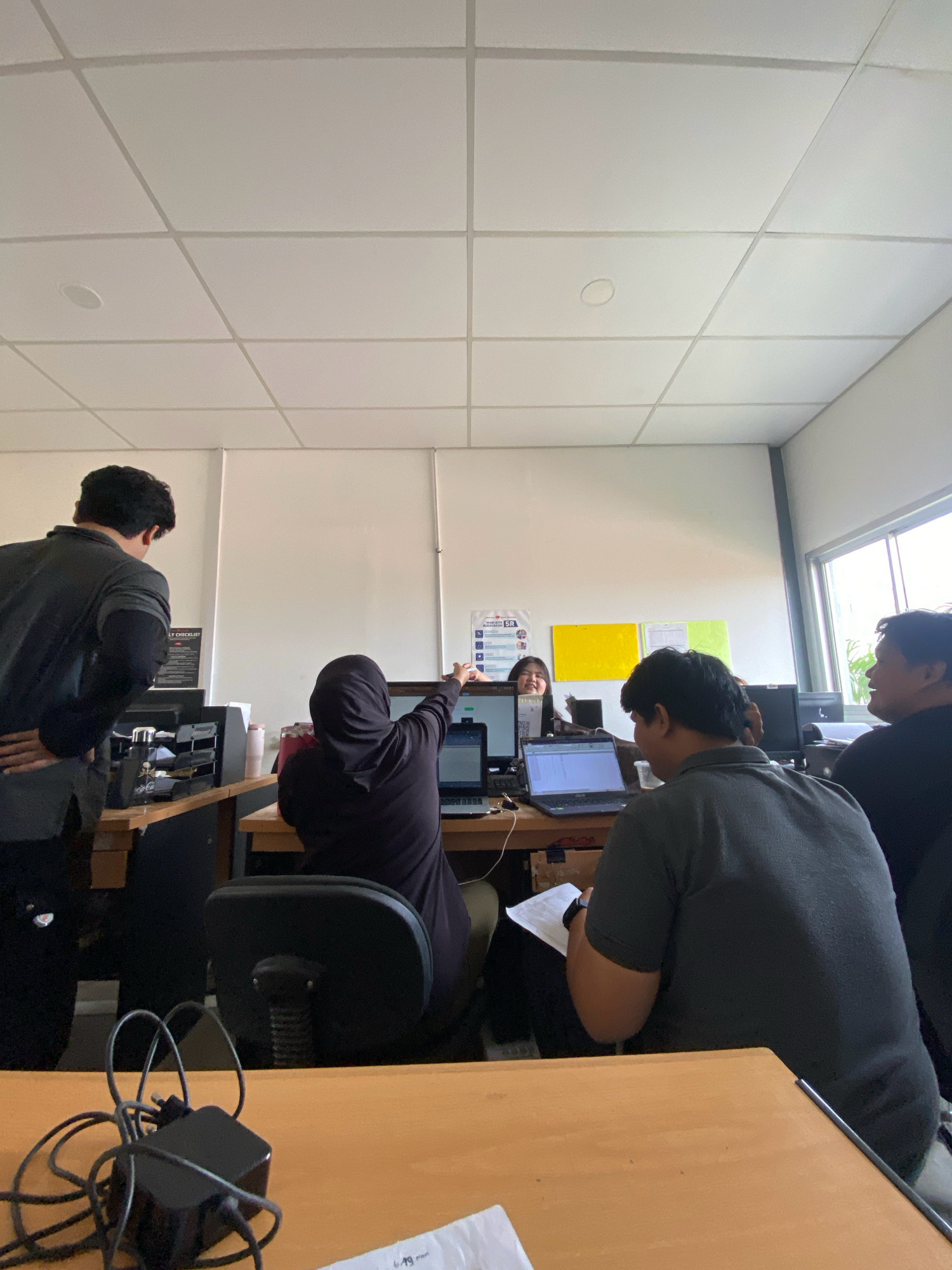 In Batam, Indonesia, four individuals are present in an office environment. One person, wearing a black hijab and outfit, is seated at a desk, pointing at a laptop screen. Another person, seen from behind and wearing a dark grey t-shirt, is seated at a desk, looking down at a piece of paper. To the left, a person in a black jacket is standing and observing the scene. To the right, a person in a black shirt is seated at a desk, looking towards the right side of the frame. The office has a white tiled ceiling with visible grid lines and a circular light fixture. Desks with computers and office supplies are arranged in the space, and a window is visible on the right wall. A power adapter with tangled cords is on the foreground desk.