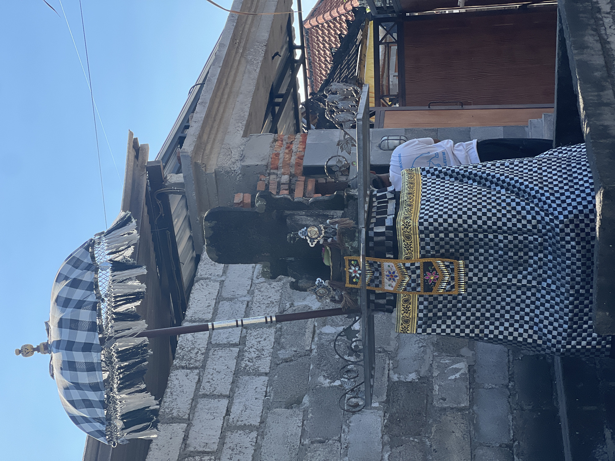 The image depicts an outdoor shrine, likely in Sukawati, Indonesia, as indicated by the architectural style and cultural elements. A tall, dark stone structure forms the central part of the shrine, adorned with intricate metalwork and fabric. A traditional Balinese umbrella, featuring a black and white checkered pattern and decorative fringe, is prominently positioned above the shrine. The shrine is draped with a black and white checkered cloth, a common element in Balinese ceremonies. In the background, a person wearing a white t-shirt and black pants stands facing away from the camera, near a wooden door. The environment appears to be a courtyard or an outdoor space with brick and concrete construction, suggesting a residential or temple setting. The overall scene conveys a sense of religious or cultural significance.