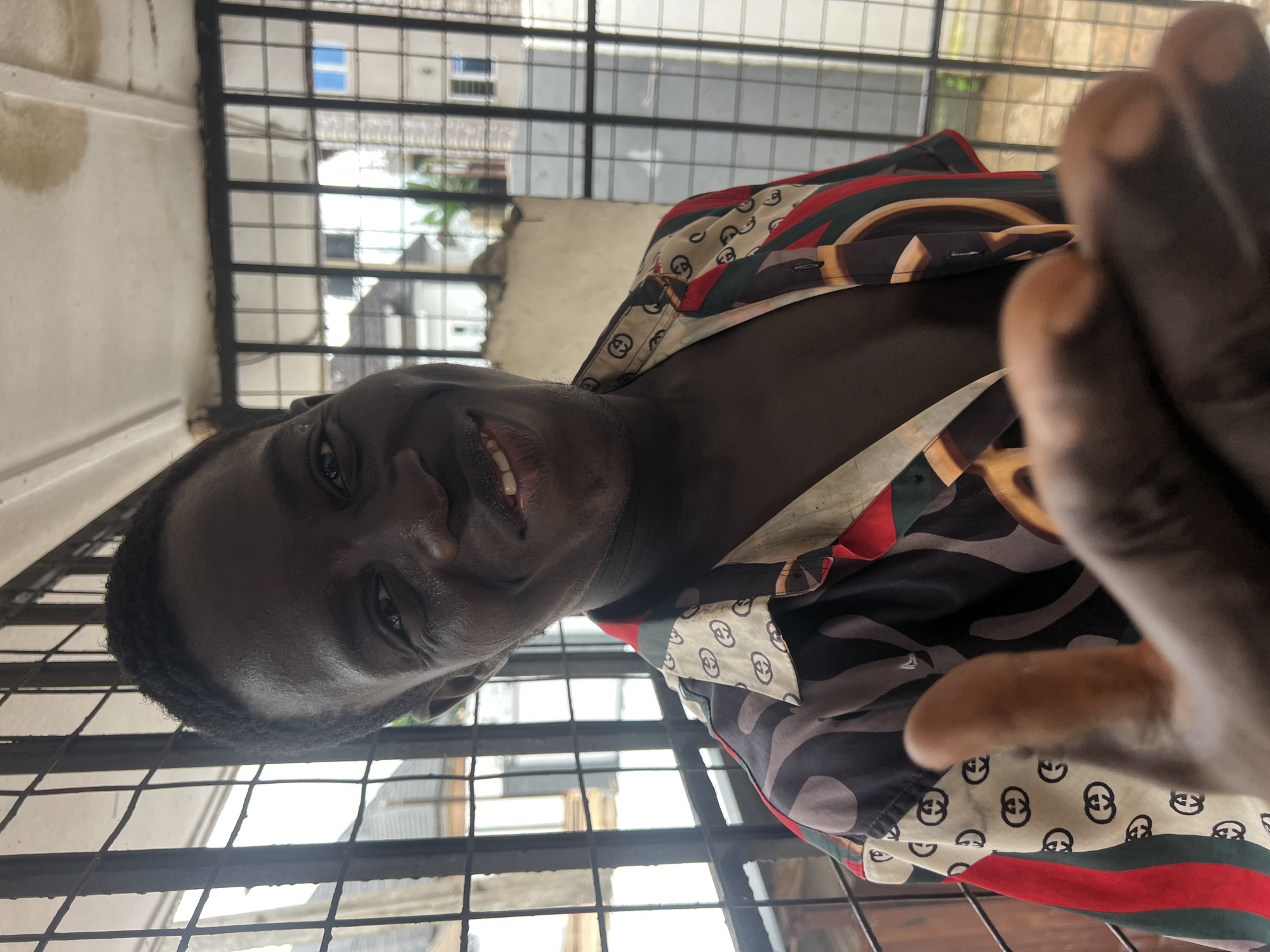 A dark-skinned man is captured in a close-up portrait, looking directly at the camera and smiling. He is wearing a colorful, patterned button-down shirt with a design that appears to be the Gucci logo repeated. The man is positioned in front of a barred window, suggesting he might be indoors or on a balcony. Through the bars, the background reveals a typical urban environment in Nkpor, Nigeria, with residential buildings visible. One building features blue-tinted windows, and another shows a lighter colored facade. The lighting in the image appears natural, indicating it was taken during the daytime. The man's right hand is prominently in the foreground, with his thumb extended towards the camera, creating a somewhat abstract and personal feel to the photograph. The overall impression is a candid and friendly snapshot, likely taken casually.