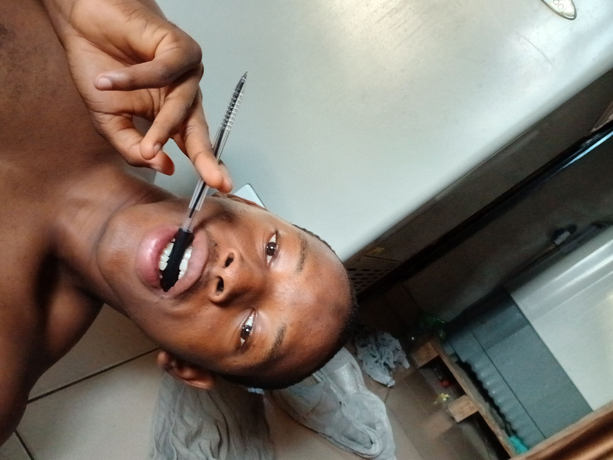 A low-angle, color photograph shows a dark-skinned person holding a pen in their mouth. The person's head is tilted, revealing an open mouth with teeth. The pen is positioned vertically in the mouth, held by the person's hand. The person's face is visible, with eyes looking towards the camera. The background includes a light-colored refrigerator, a wooden structure, and what appears to be discarded cloth. The location is presumed to be Lagos, Nigeria.