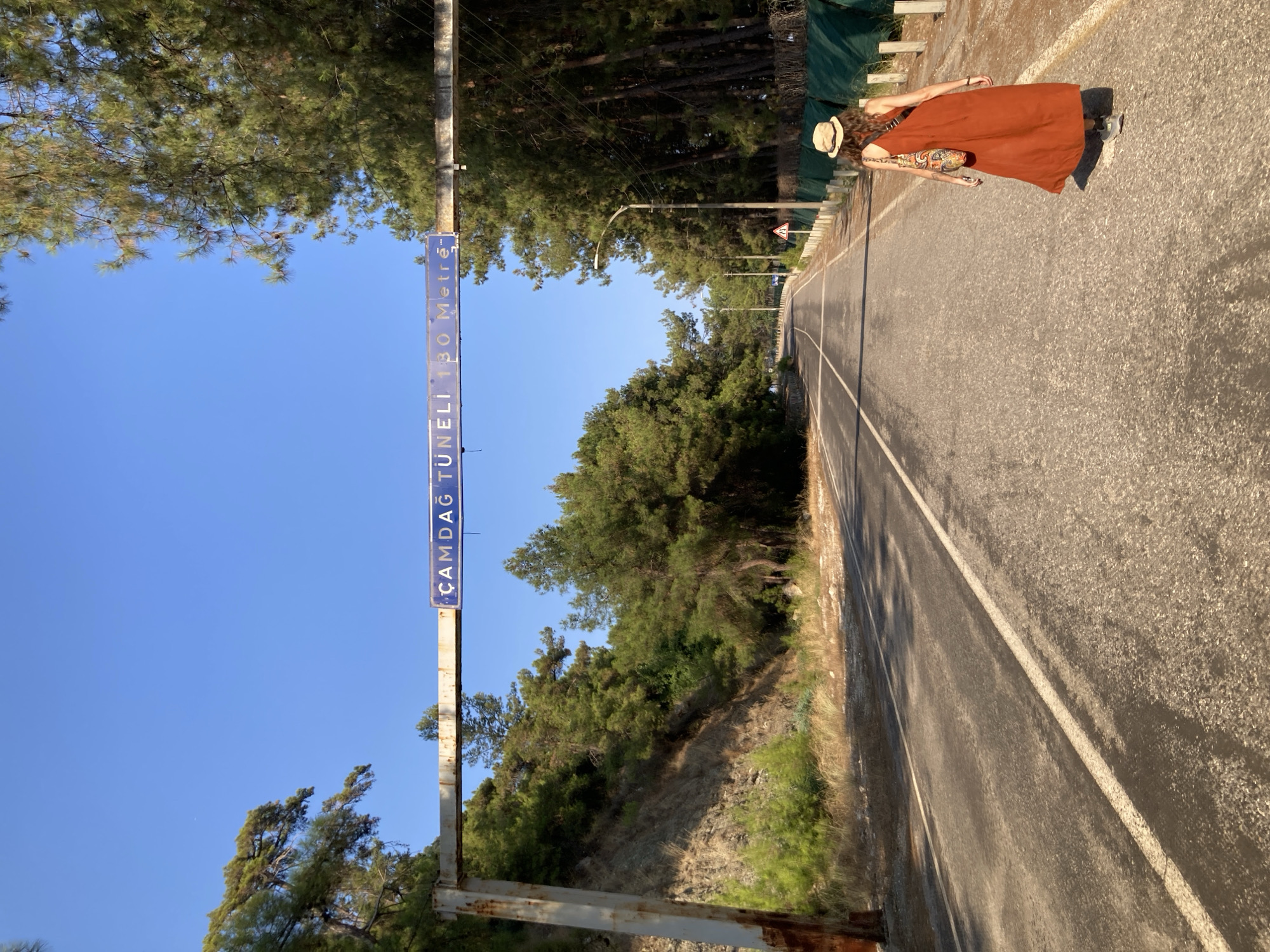 A woman walks away from the camera down a paved road lined with pine trees under a clear blue sky. She wears an orange dress and a straw hat, with a patterned bag slung over her shoulder. A blue sign overhead reads "ÇAMDAĞ TUNELİ 130 Metre". The road leads into a forested area where a tunnel is indicated by the sign. The location is Kemer, Turkey.