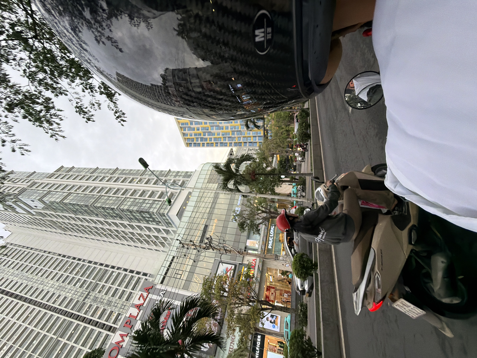 The image captures a street scene in Nha Trang, Vietnam, from the perspective of someone riding on a scooter. In the foreground, the shiny black helmet of the rider is prominent, reflecting the surrounding environment, including buildings and trees. A mirror on the scooter's handlebars shows a reflection of a person wearing a white shirt. In the middle ground, another person is riding a beige scooter, wearing a red helmet and a gray hoodie. They are on the right side of a multi-lane road. Cars and other scooters are visible on the road. In the background, a very tall, modern white building with many windows dominates the skyline, with "OM PLAZA" visible on its facade. Another colorful, modern building with yellow and blue accents is also present. Palm trees and other foliage are scattered throughout the scene, indicating a tropical or subtropical urban environment. The sky is overcast, suggesting it might be early morning or late afternoon. The overall impression is of a busy urban street in Nha Trang.