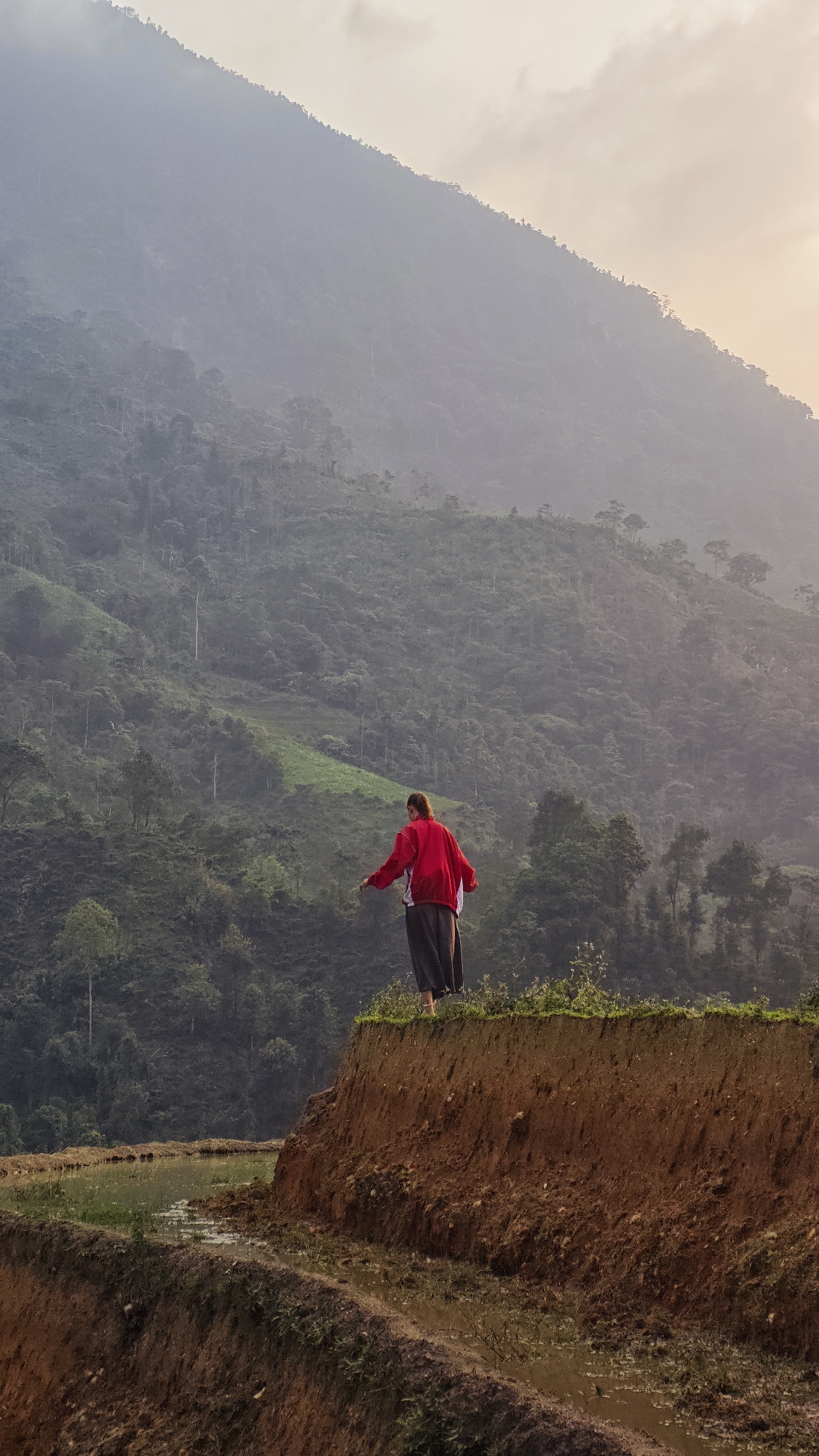 A woman stands at the edge of a terraced field, gazing out at a sprawling, mist-shrouded mountainous landscape in Nậm Dịch Commune, Vietnam. She is positioned with her back partially to the viewer, wearing a vibrant red jacket or sweater and dark, flowing lower garments. Her arms are slightly outstretched, suggesting a moment of contemplation, balance, or admiration for the view.
The foreground features the rugged, reddish-brown earth of the steep terraced fields, with standing water visible in the lower paddies, reflecting the subtle light. Sparse green vegetation dots the immediate ground around the woman. Behind her, dense, forested mountains rise in undulating layers, fading into a soft atmospheric haze that imbues the scene with an ethereal quality. The sky above, visible towards the upper right, displays soft, warm tones, indicative of either early morning or late afternoon light. The overall mood is one of tranquility and natural grandeur. There is no visible text.