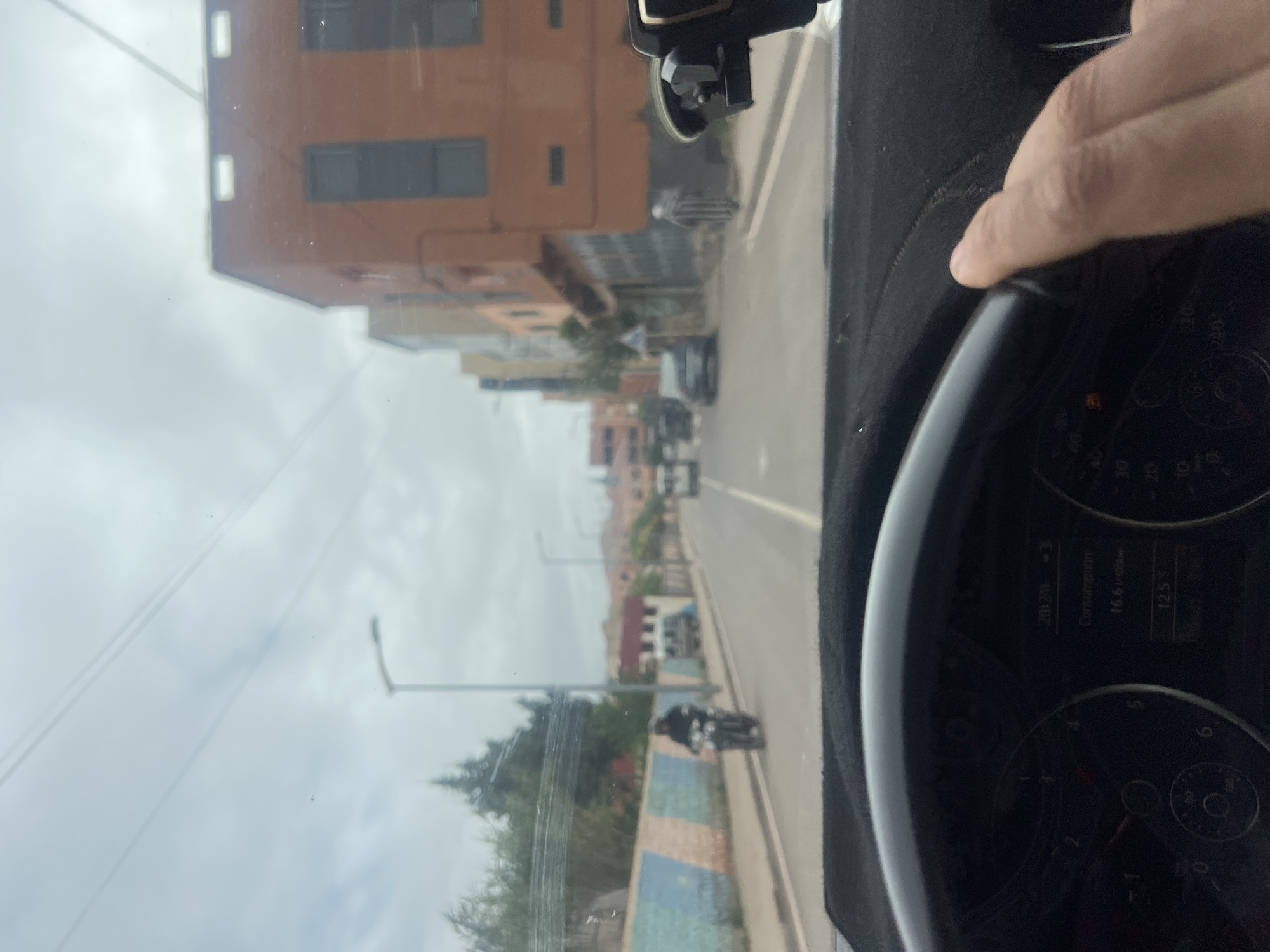 The view is from inside a vehicle, presumably a car, as suggested by the dashboard components visible on the right side of the frame. The hand of the driver is visible resting on the dashboard. Looking through the windshield, the scene depicts a street in Ouaouizert, Morocco.
The buildings along the street are predominantly brown and orange. The sky is overcast. Various vehicles are visible on the road. A motorbike is also visible in the foreground. The street is lined with streetlights and trees.