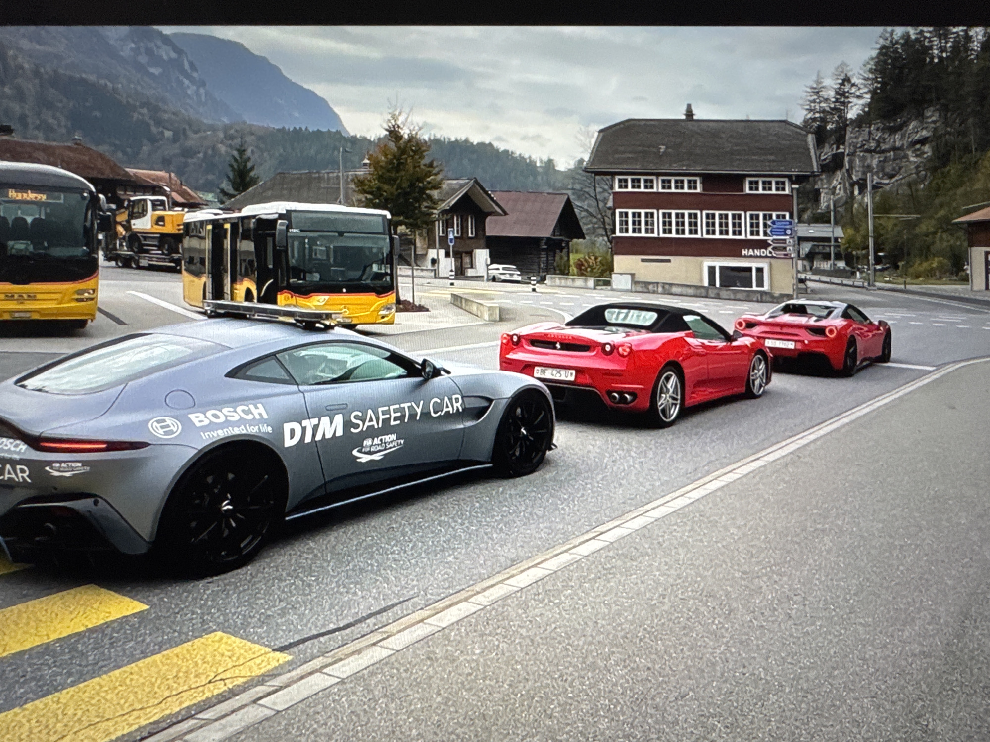 The image shows a street scene in what appears to be a town with a mountainous backdrop. In the foreground, a gray safety car with "DTM SAFETY CAR" written on its side is parked. Behind it, two red sports cars are visible. Further back, two yellow buses and a construction vehicle are parked on the side of the road, near some buildings that resemble traditional Swiss architecture. The sky is overcast, suggesting a cloudy day. The location is identified as Porrentruy, Switzerland.
